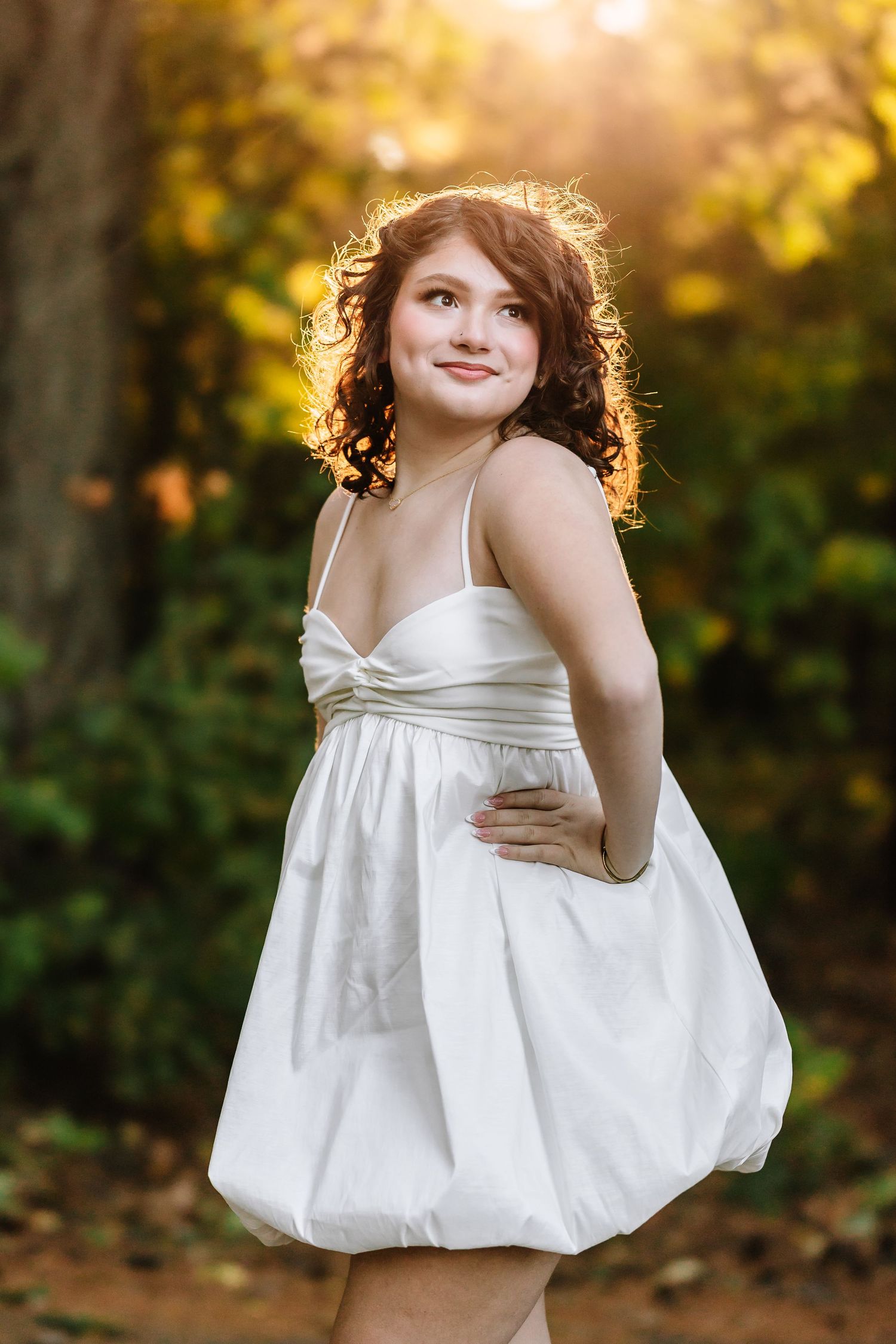 Young woman in white bubble dress smiling outdoors in golden autumn light among colorful trees.