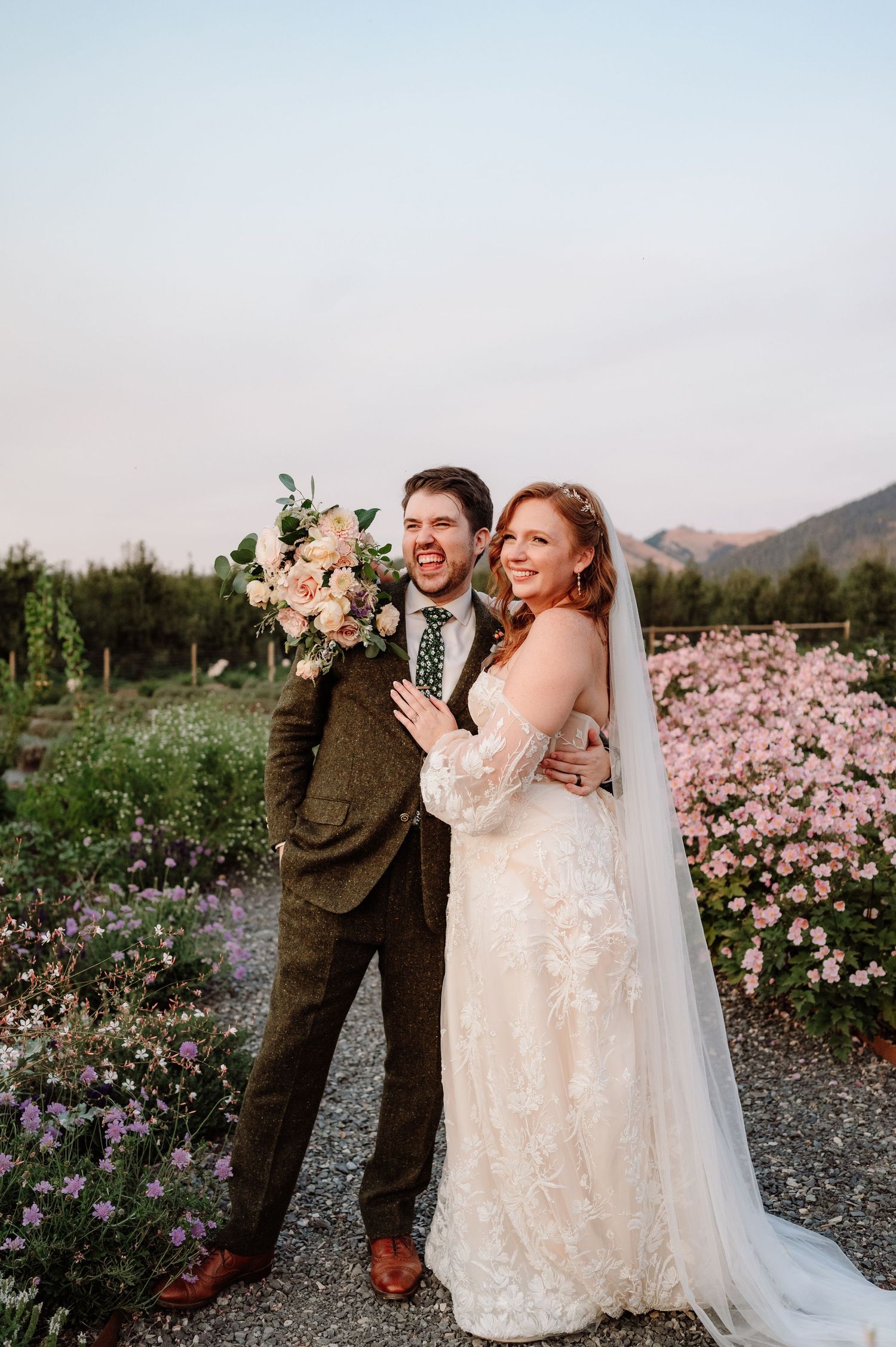 Joyful bride and groom pose playfully among pink flowers with mountain views at dusk.