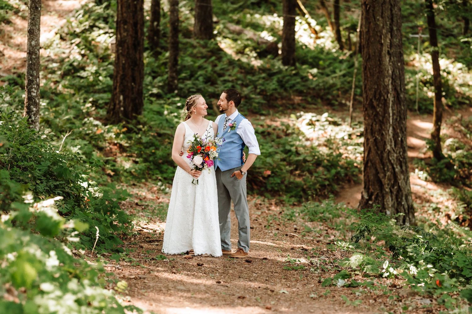 Bride and groom share a tender moment on a forest path surrounded by tall pine trees.