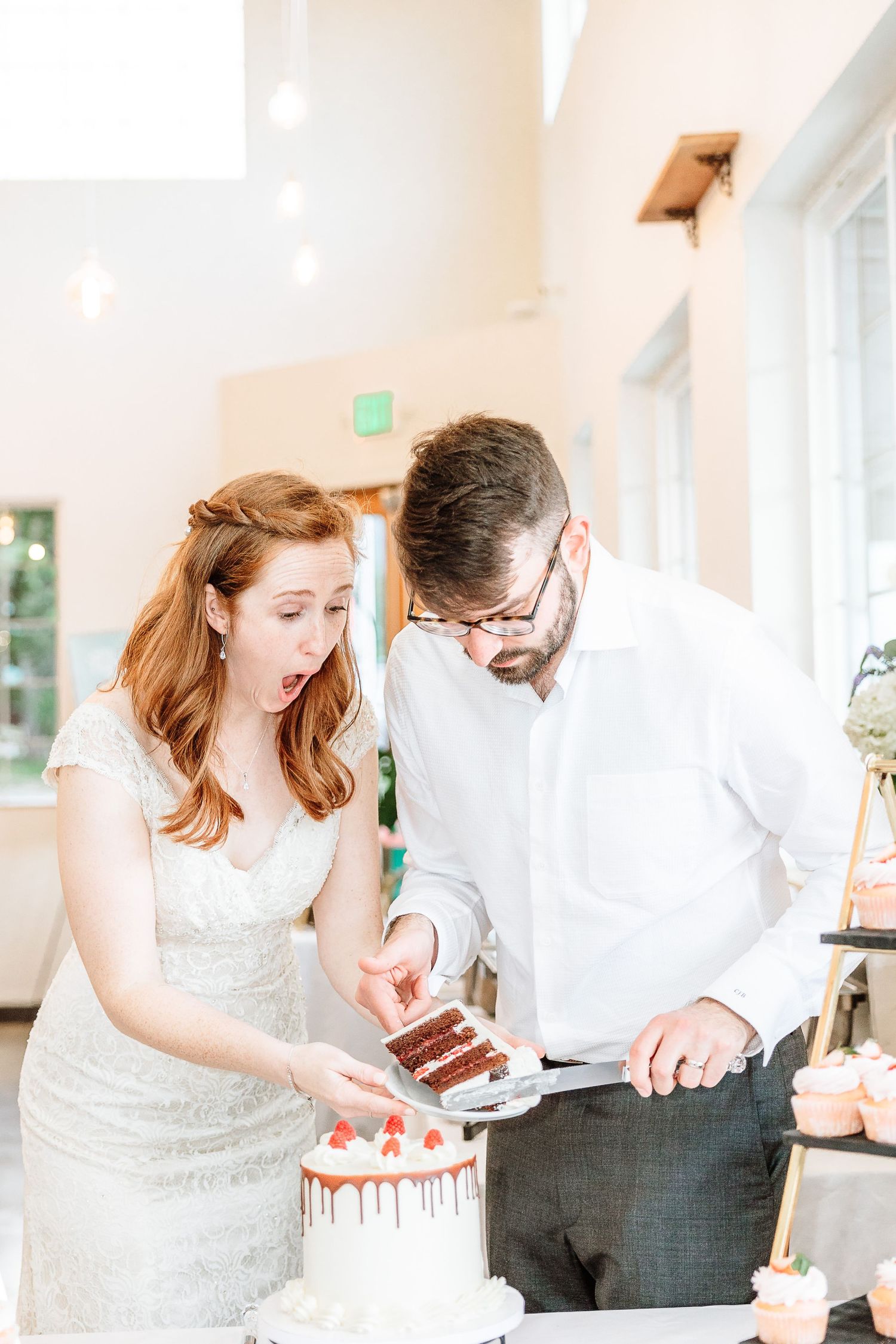 Bride reacts with surprise as groom serves wedding cake slice at bright modern reception venue.
