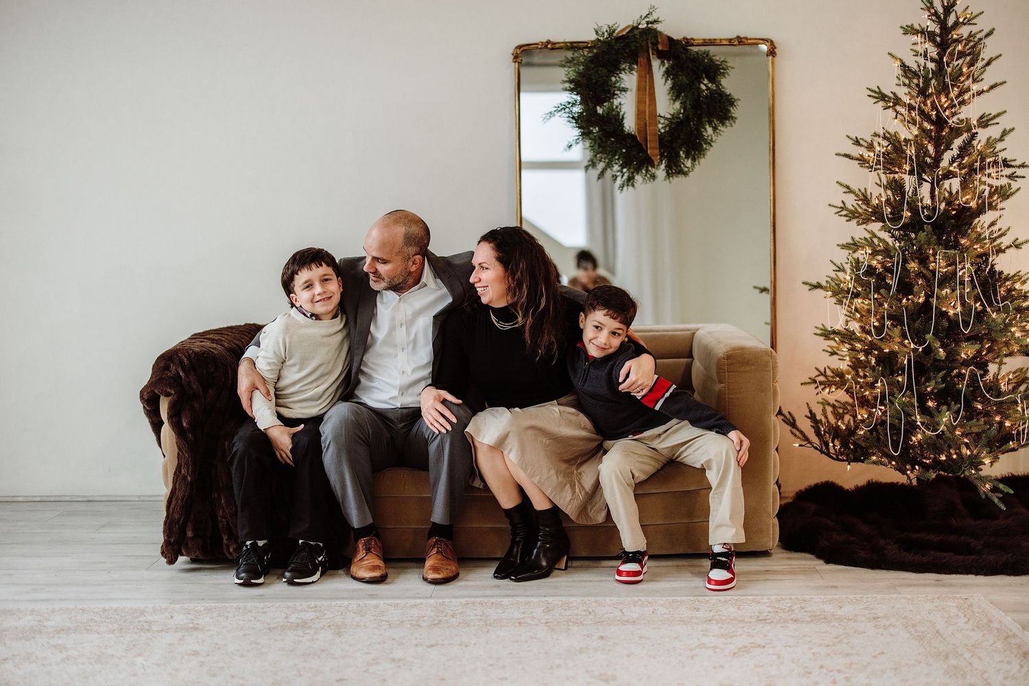 Family of four sitting on a couch by a Christmas tree in a festive holiday portrait session.