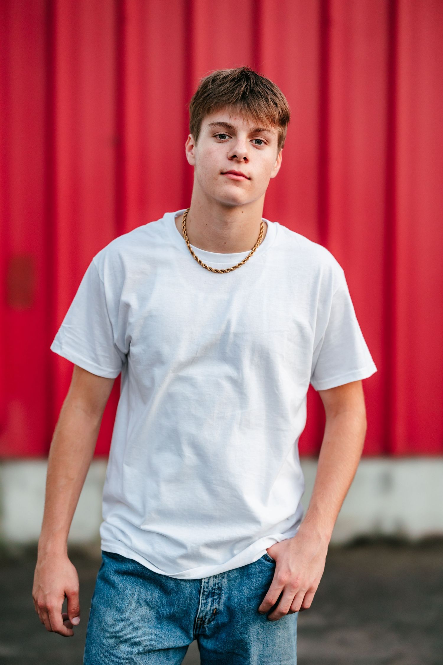 Young man in white t-shirt and gold chain necklace posing against a red wall wearing blue jeans.