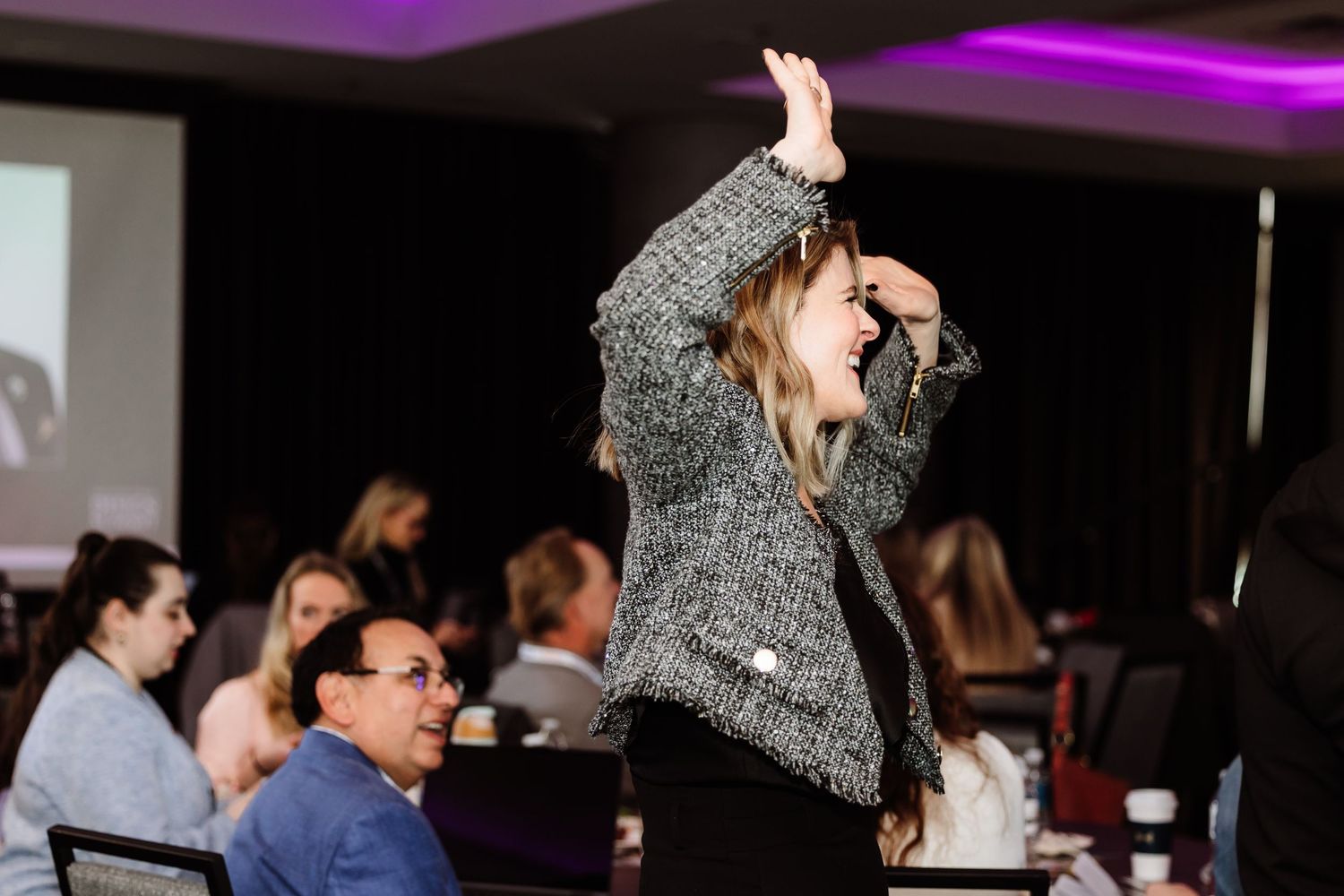 An enthusiastic woman raises her hand laughing during a conference session.