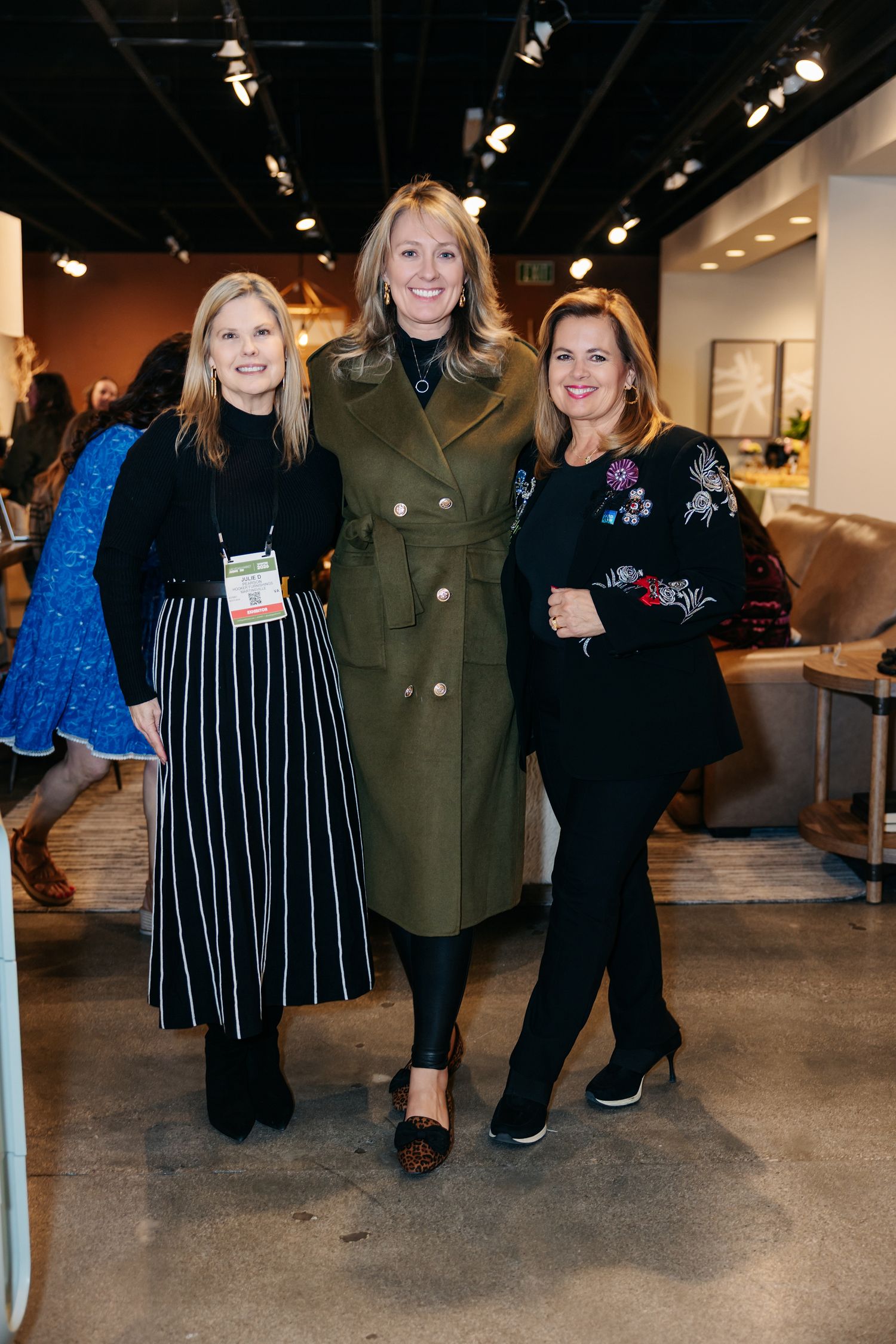 Three women pose together smiling at a furniture trade show event.