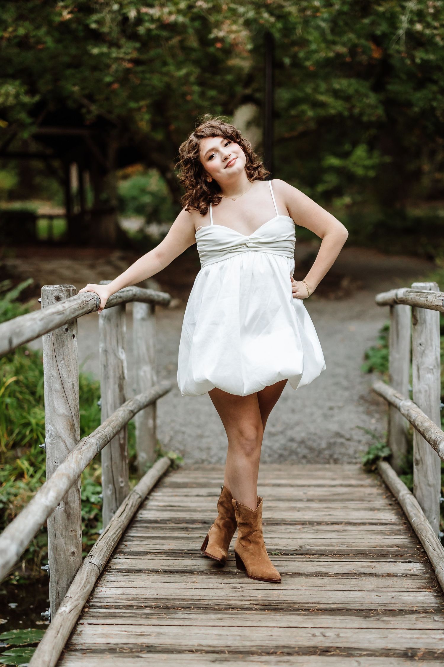 Young woman in white bubble dress and brown cowboy boots posing on rustic wooden bridge surrounded by lush greenery.