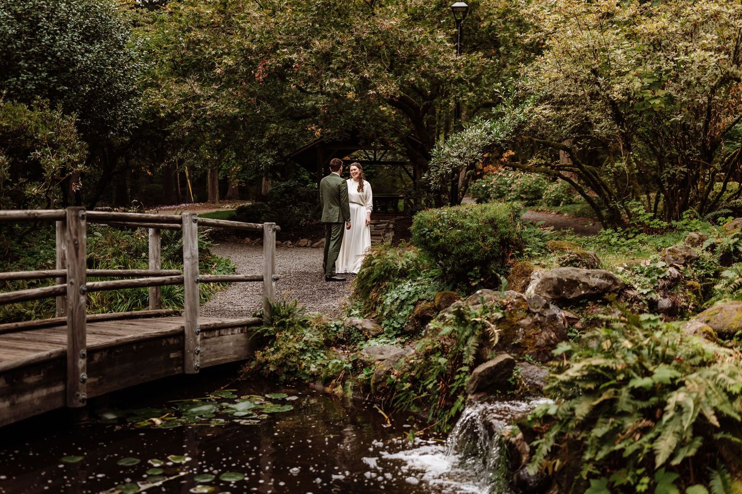 Couple in wedding attire stands on a stone path by a wooden bridge in a lush Japanese garden with a small waterfall.