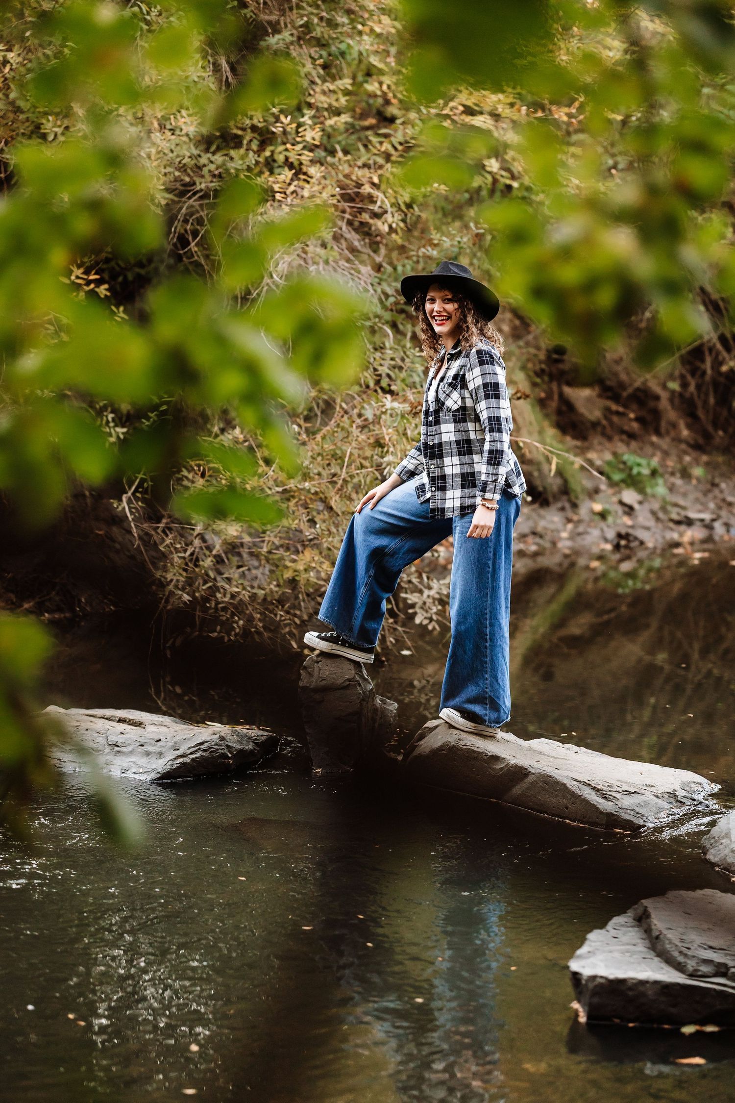 Woman in plaid shirt, jeans, and cowboy hat standing on rocks by a creek surrounded by lush greenery.
