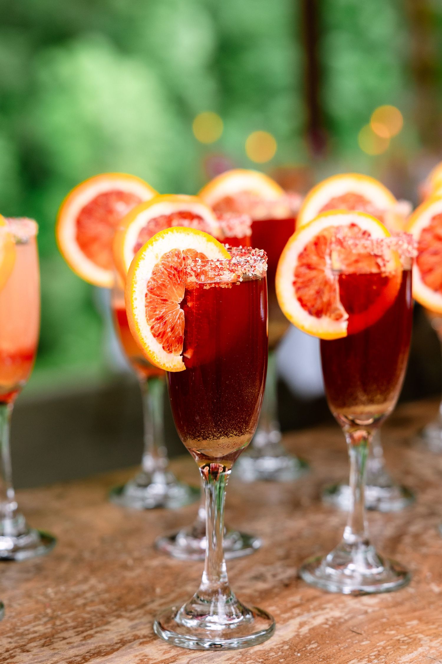 Row of deep red blood orange mimosas with sugared rims and citrus garnishes on a wooden bar.