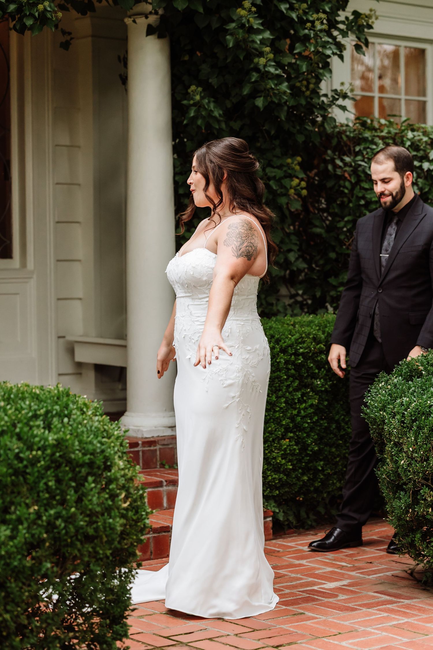 Bride in strapless lace wedding gown with tattoo smiling as groom approaches outdoors.