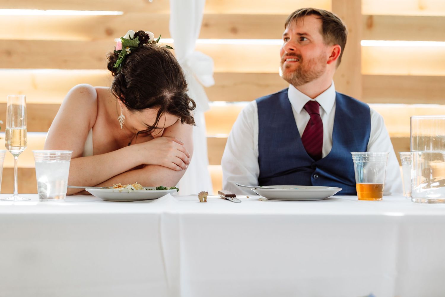 Bride laughing with head down at wedding reception table while groom smiles beside her.