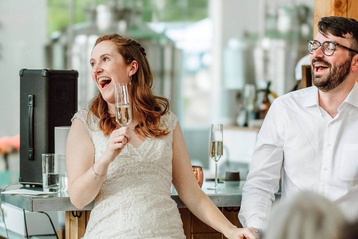 Bride and groom laugh joyfully while holding champagne glasses at casual brewery wedding reception.