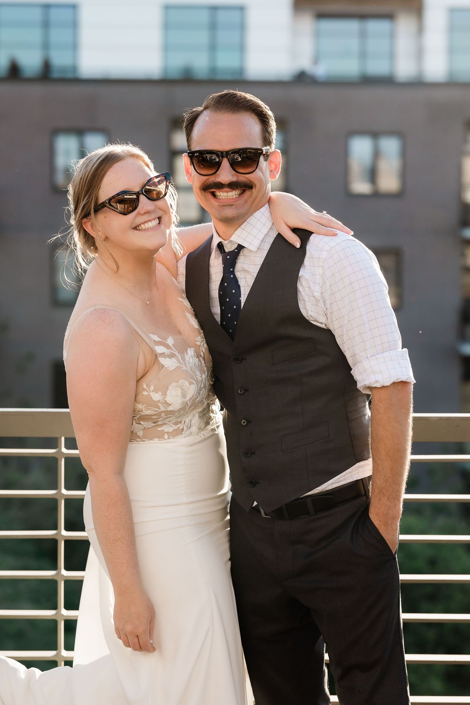 Bride and groom pose together wearing sunglasses on sunny outdoor balcony.