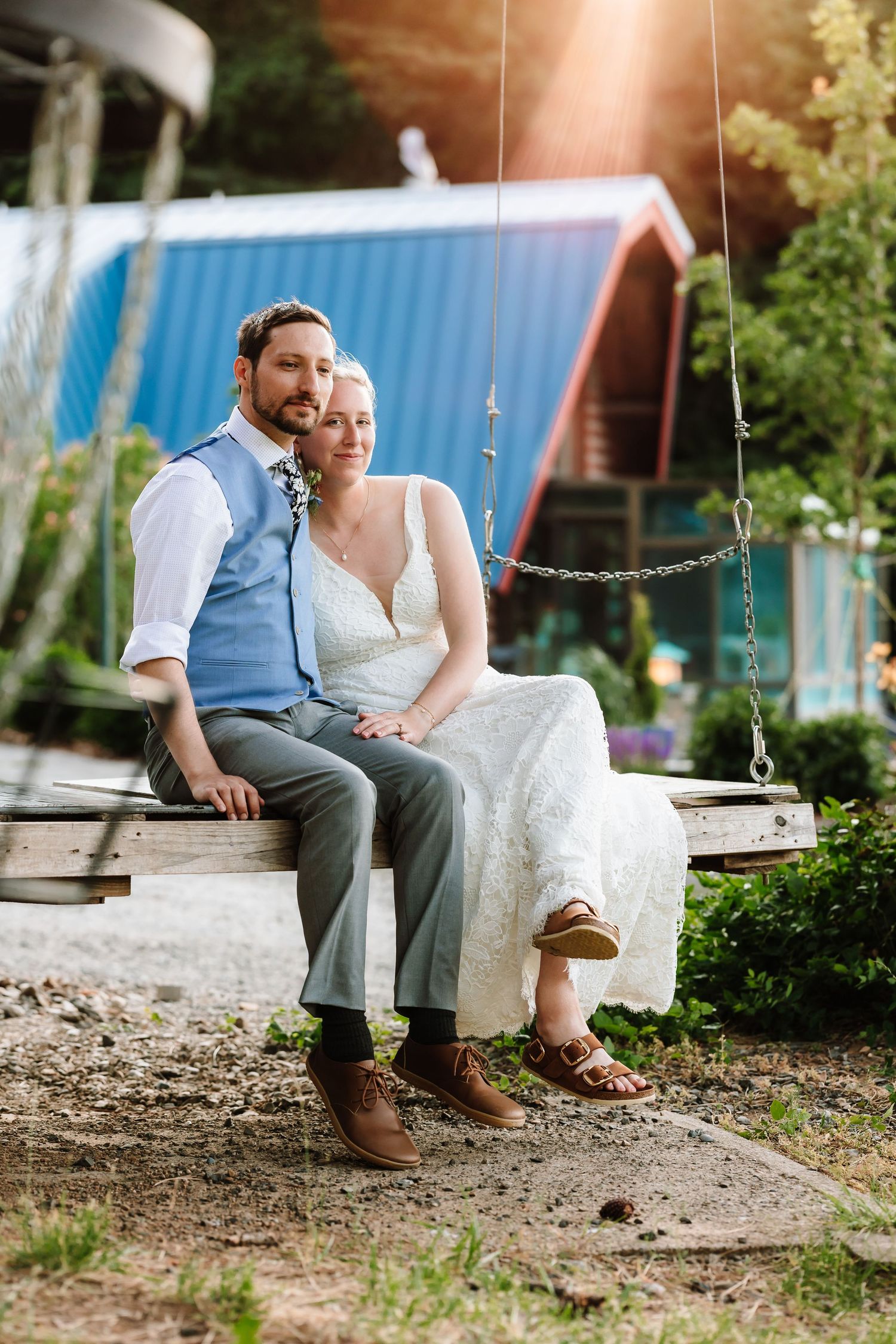 Bride and groom sit together on a wooden swing in golden sunlight near a blue building.