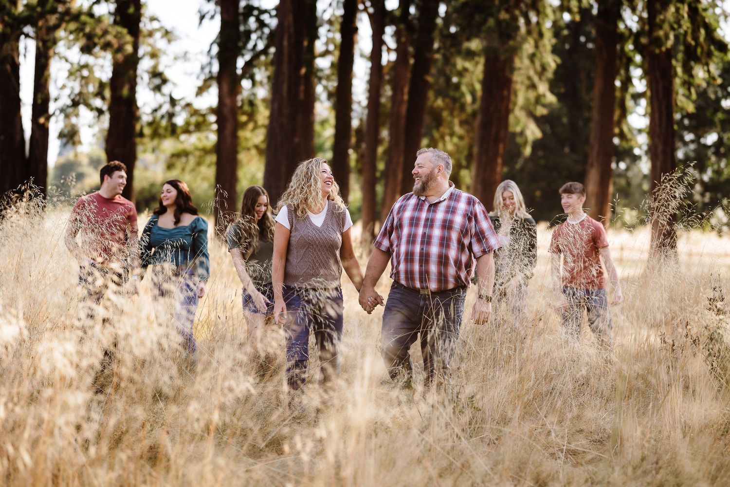 A family walks hand-in-hand through a golden field surrounded by tall pine trees in warm sunlight.