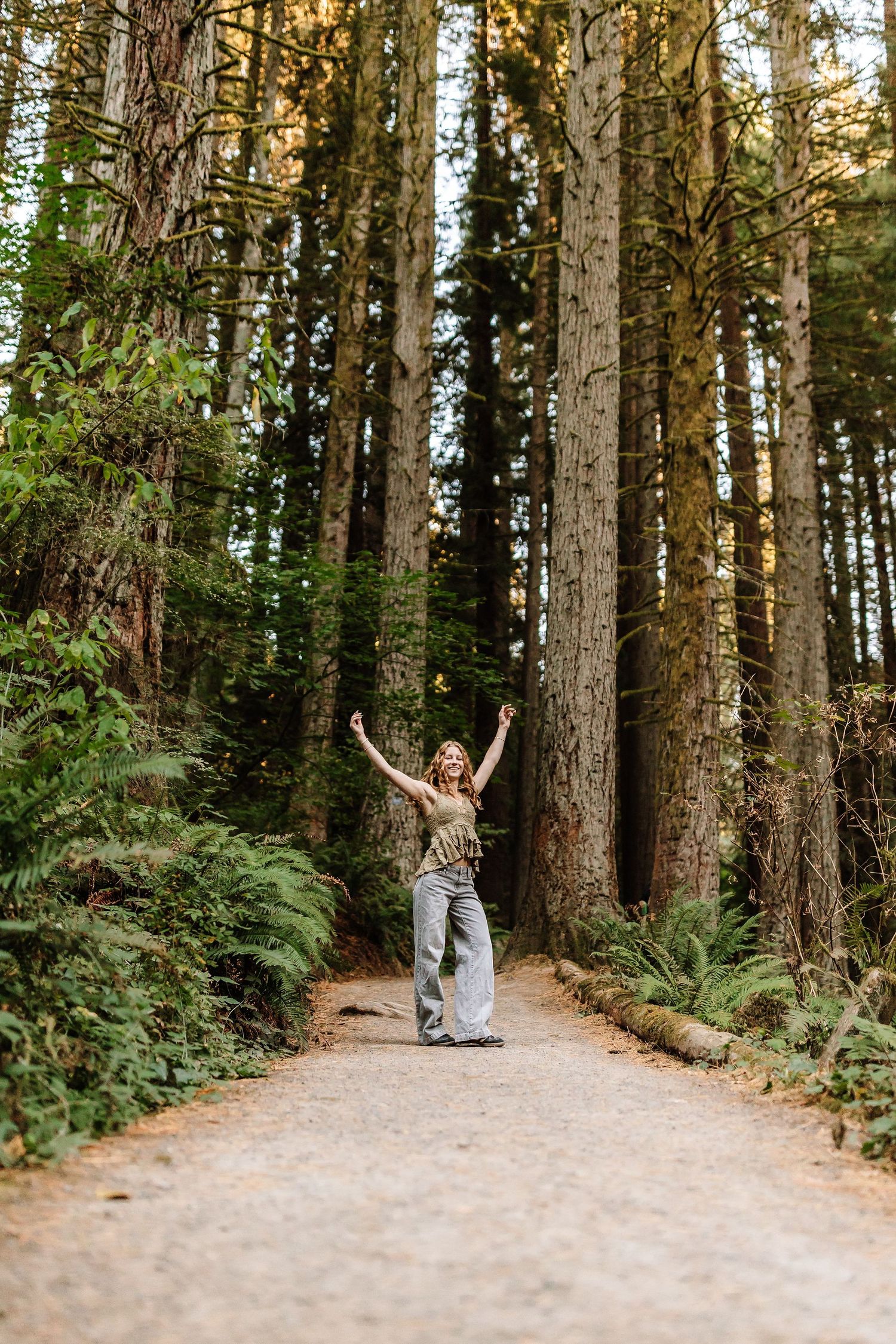 Woman with arms raised joyfully on a forest path surrounded by tall trees and ferns.