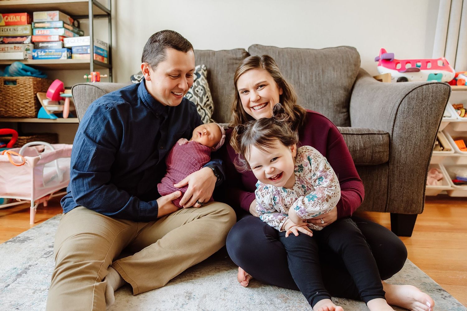 Smiling family of four sitting on living room floor with newborn and toddler.