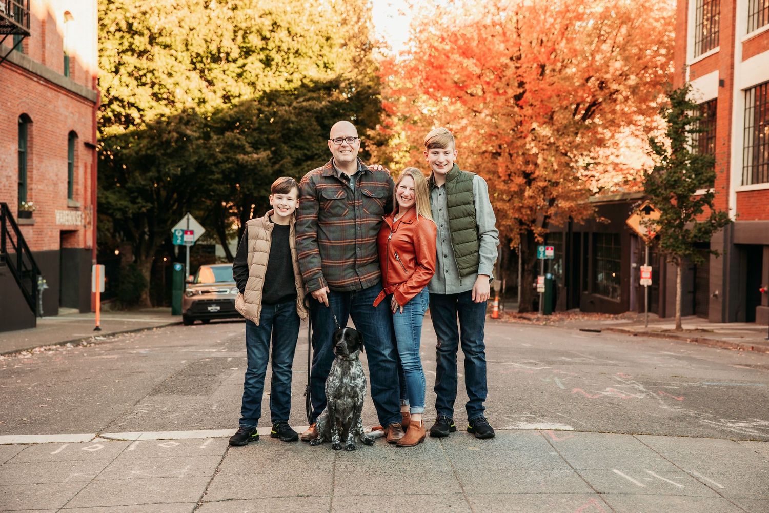Family of four with a dog posing on a brick street surrounded by vibrant fall foliage in an urban setting.