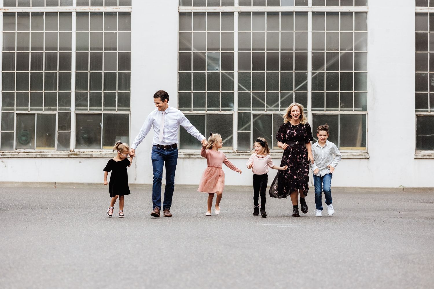 A family of six holds hands and walks together in front of large industrial windows outdoors.