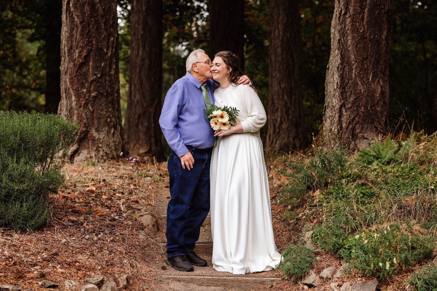 An older man kisses a bride on the cheek as she holds a bouquet in a lush forest setting.