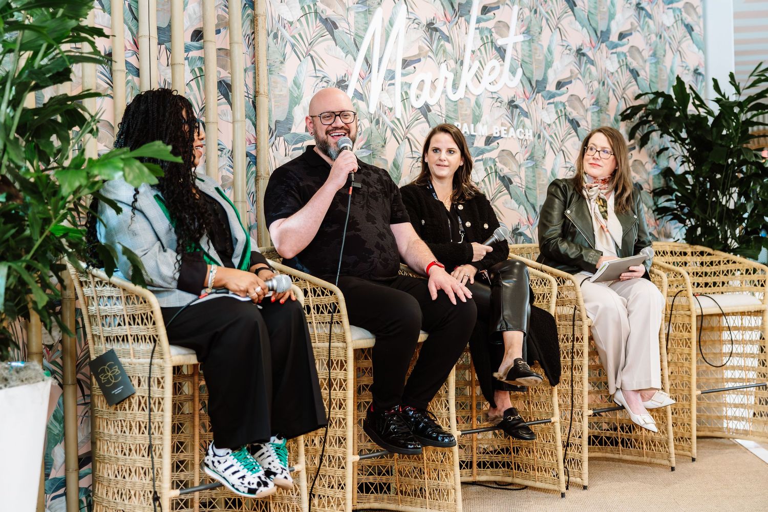 Four panelists in rattan chairs at tropical-themed market discussion event with greenery backdrop.