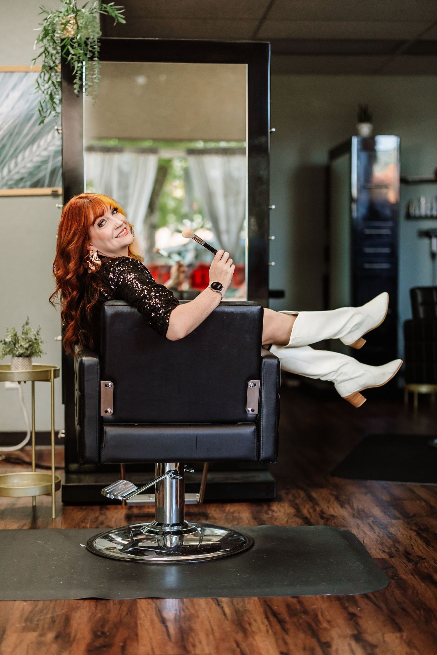 Redhead woman in black dress and white boots relaxing in salon chair holding phone.