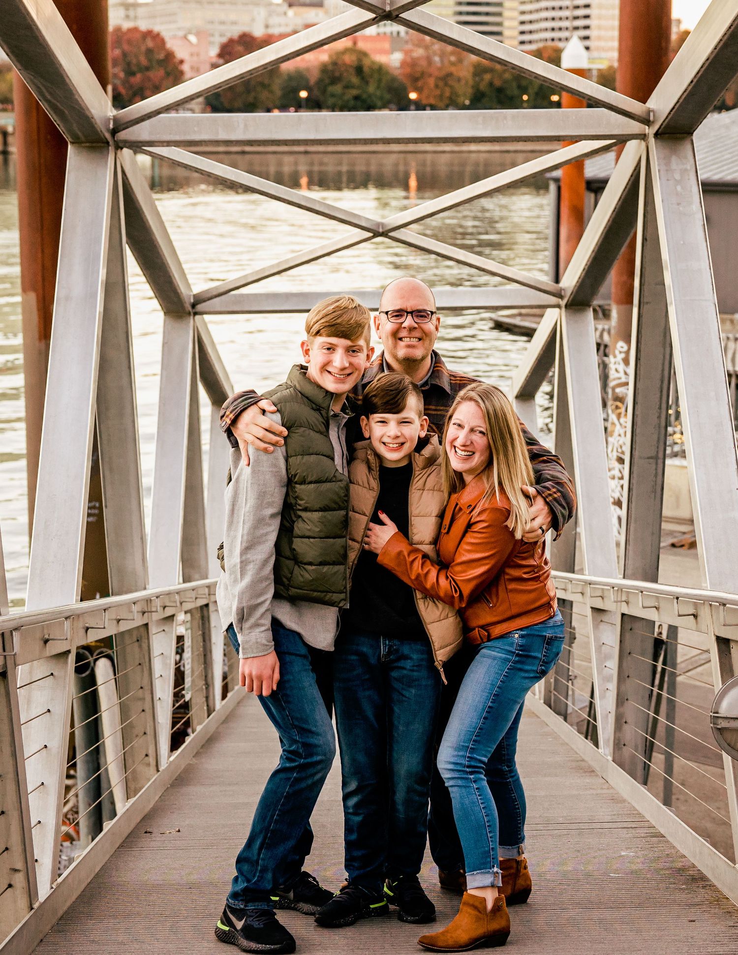 Family of four smiling together on a bridge walkway with a river in the background during fall season.