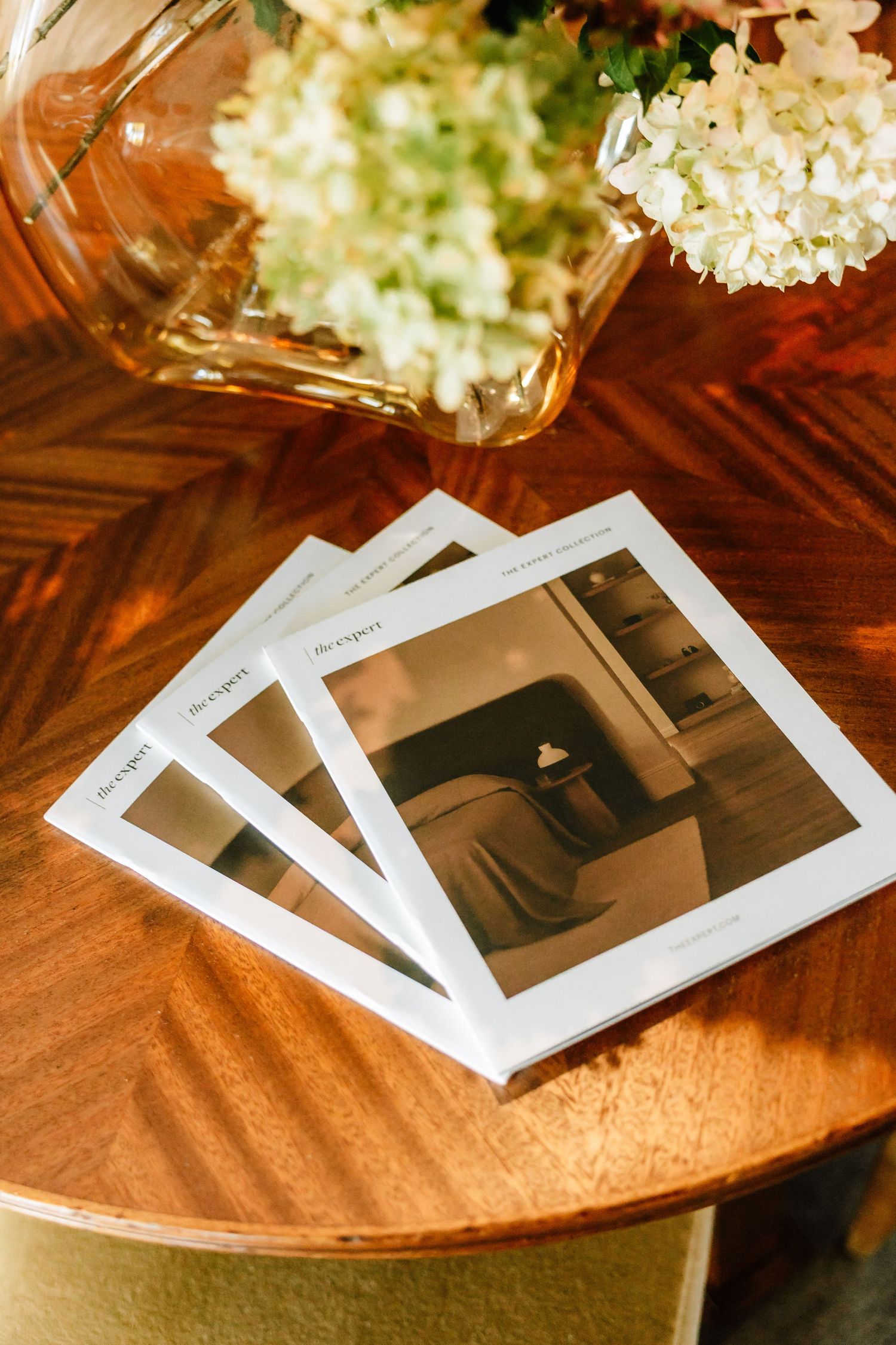 Three design catalogs stacked on wooden table beside fresh white hydrangea floral arrangement.