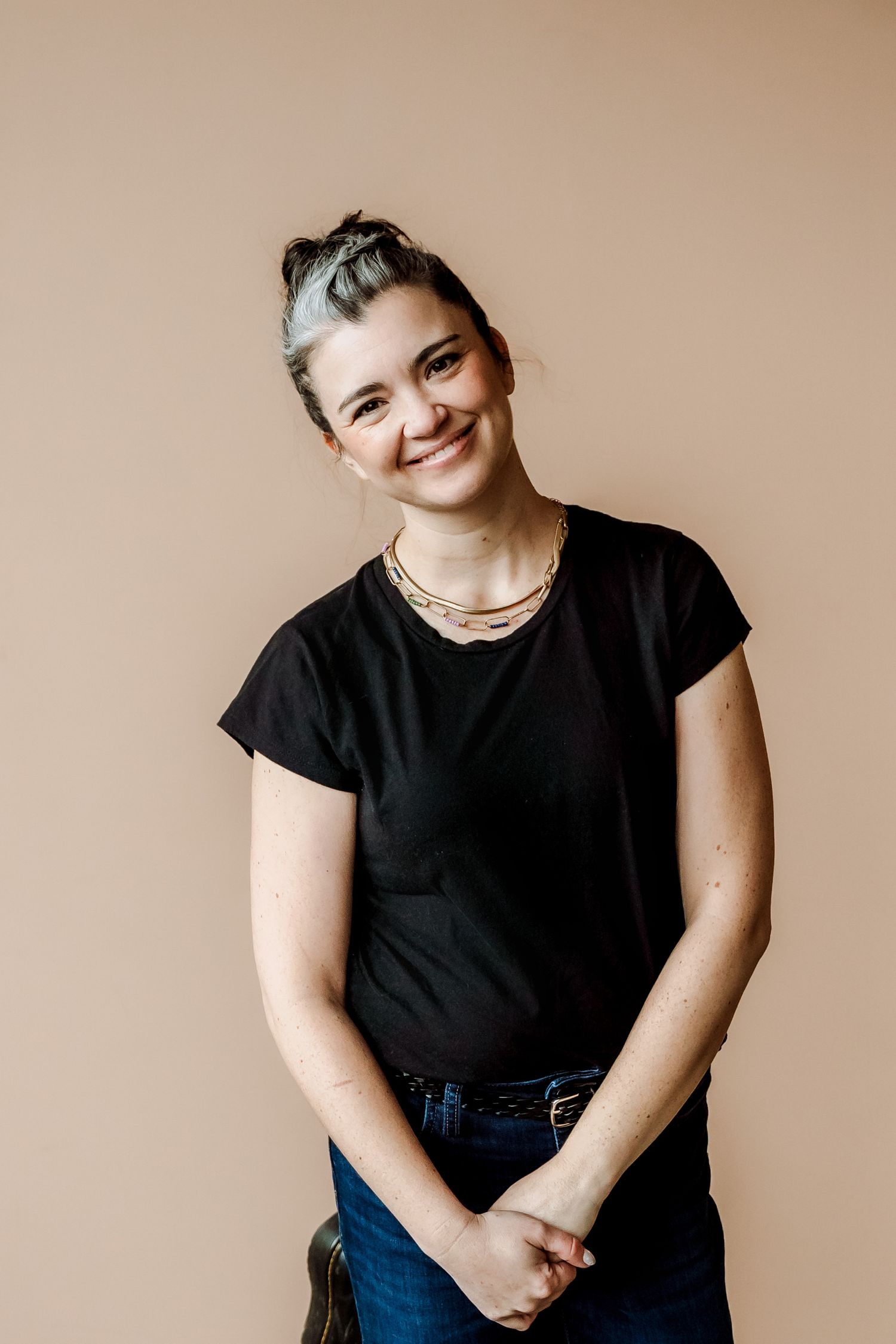 Smiling woman with gray-streaked updo wearing black t-shirt and gold necklace against beige background.