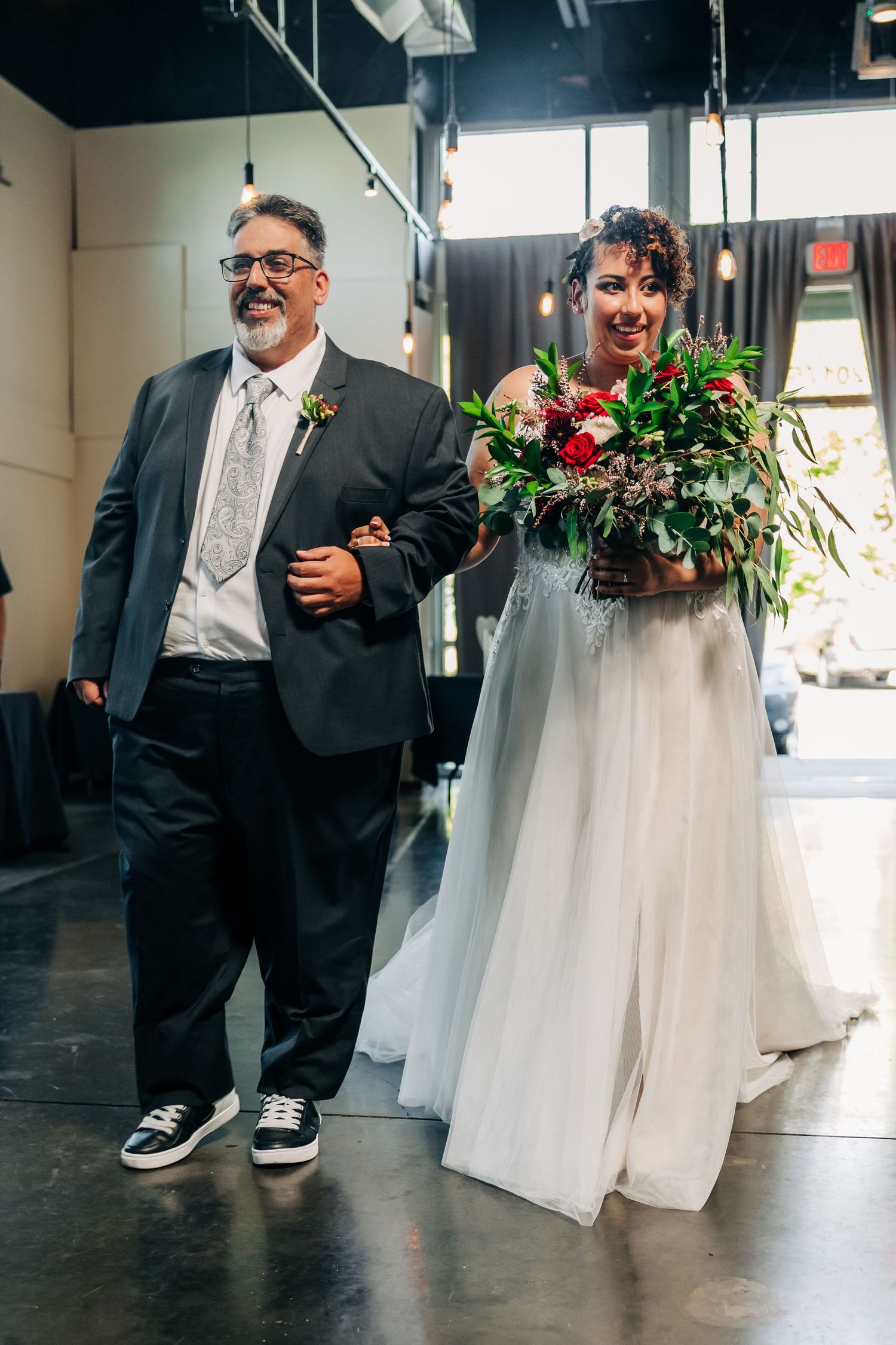 Bride carrying lush red bouquet walks down aisle with father during indoor wedding ceremony.