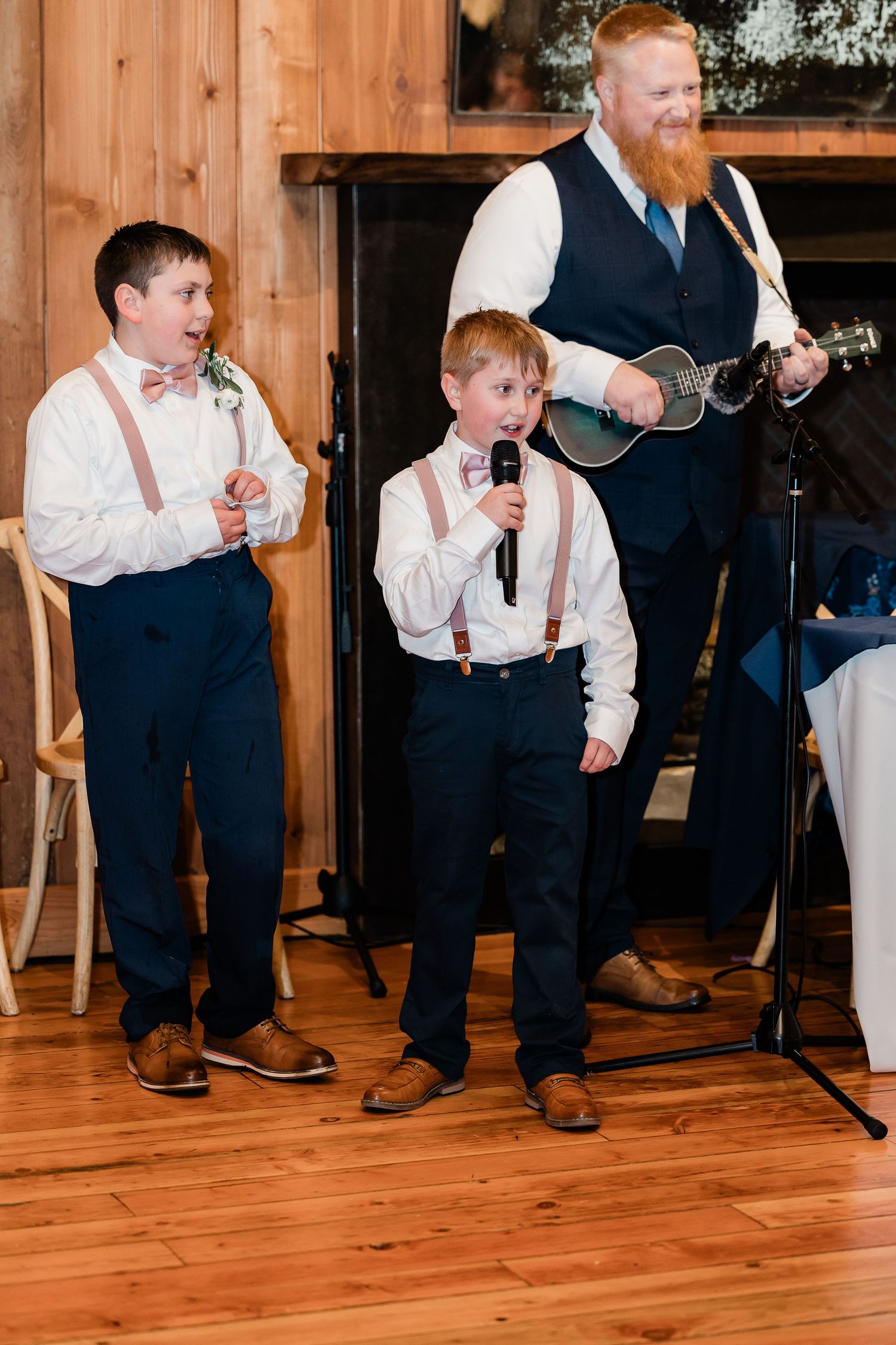 Two young boys in suspenders and bow ties sing into microphone at wedding reception with guitarist.