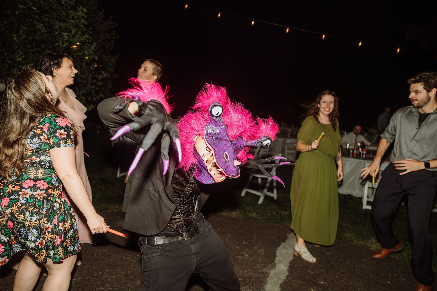 Wedding guests dance outdoors at night with a colorful pink feathered pinata costume centerpiece.