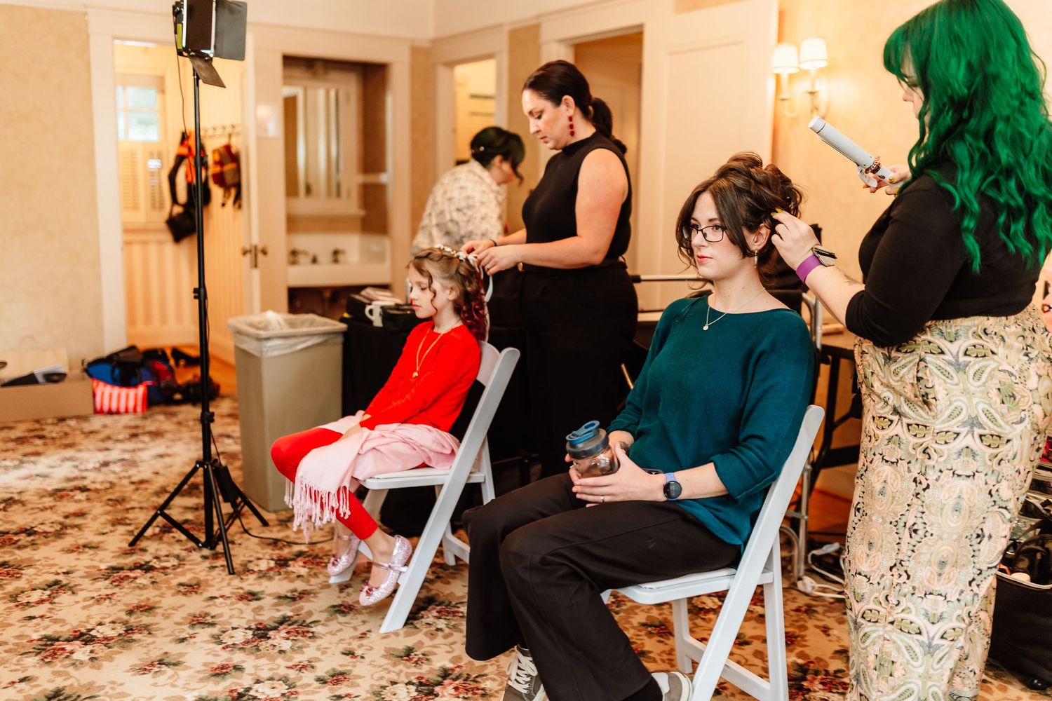 Women getting hair and makeup done in a bright room before a wedding.