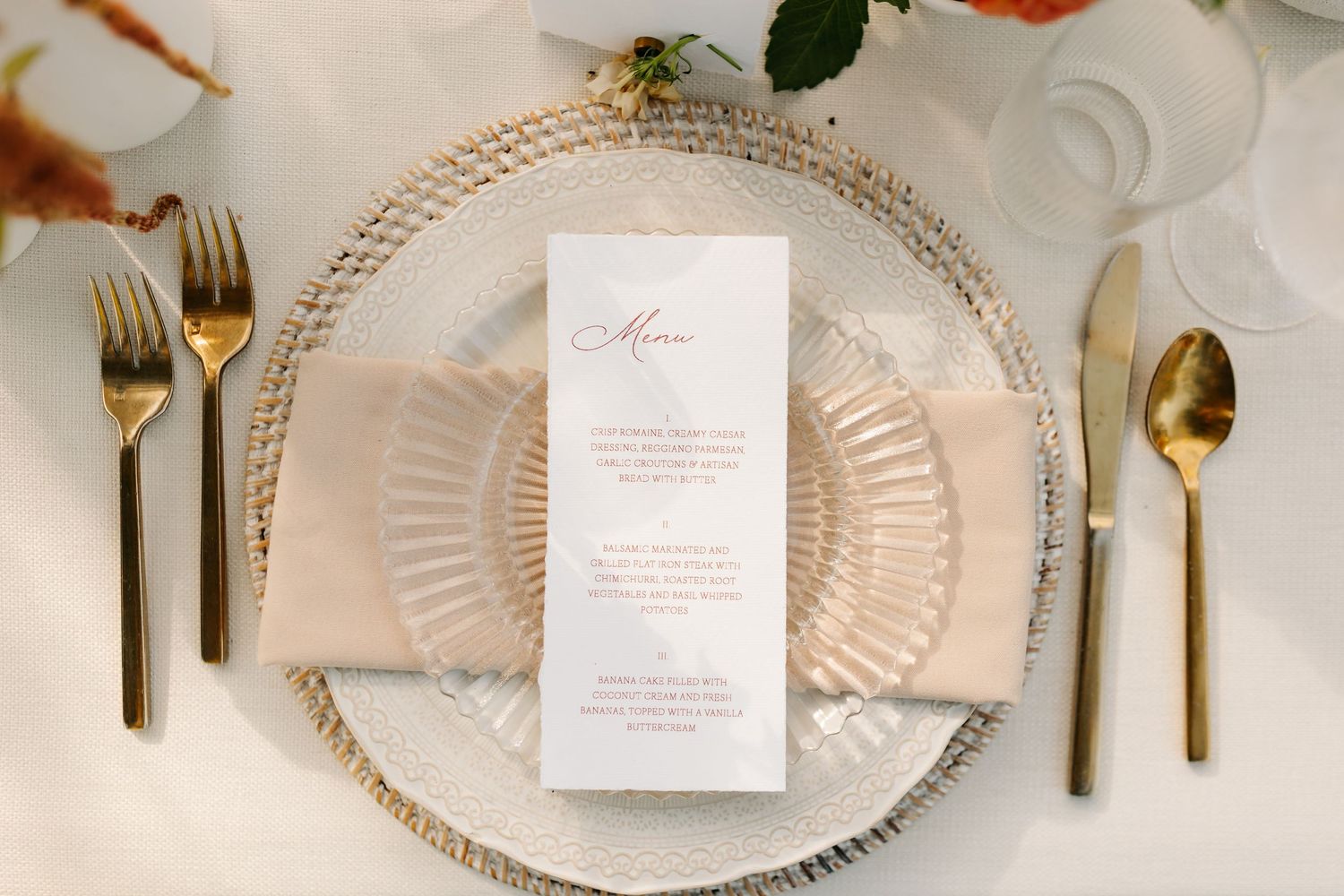 Elegant wedding place setting with glass charger, blush napkin, gold cutlery, and white menu card.