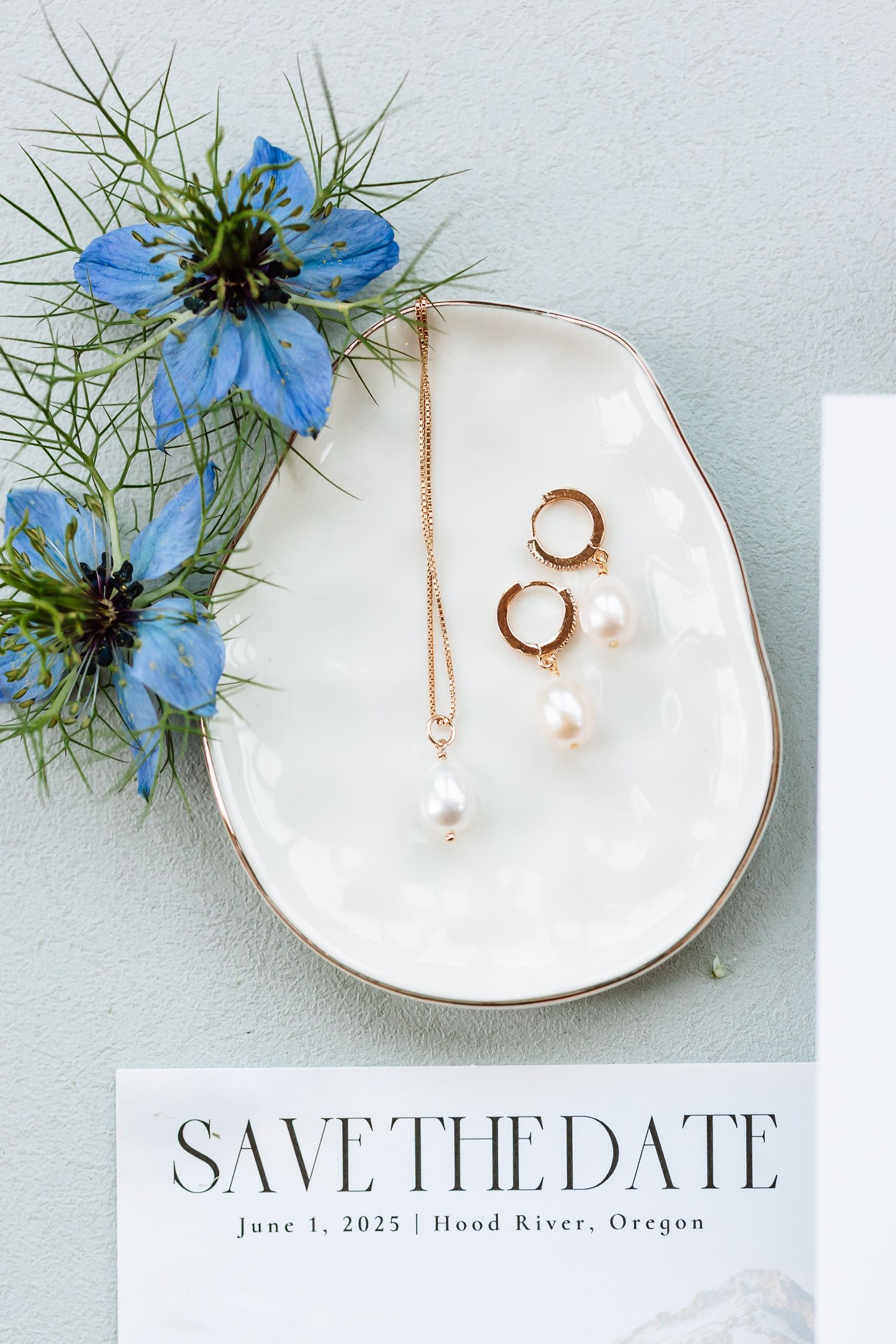 Pearl drop earrings and necklace on white ceramic dish with save the date card and blue nigella flowers.
