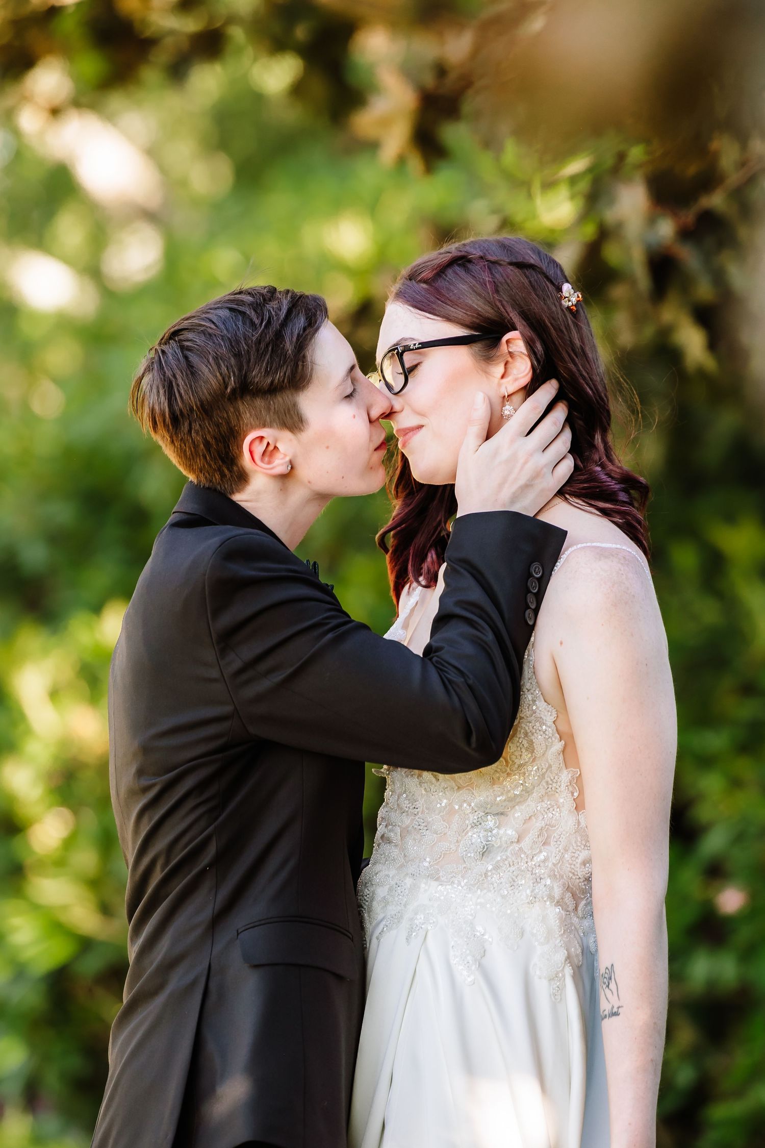 Two brides share a romantic kiss outdoors surrounded by lush green foliage.