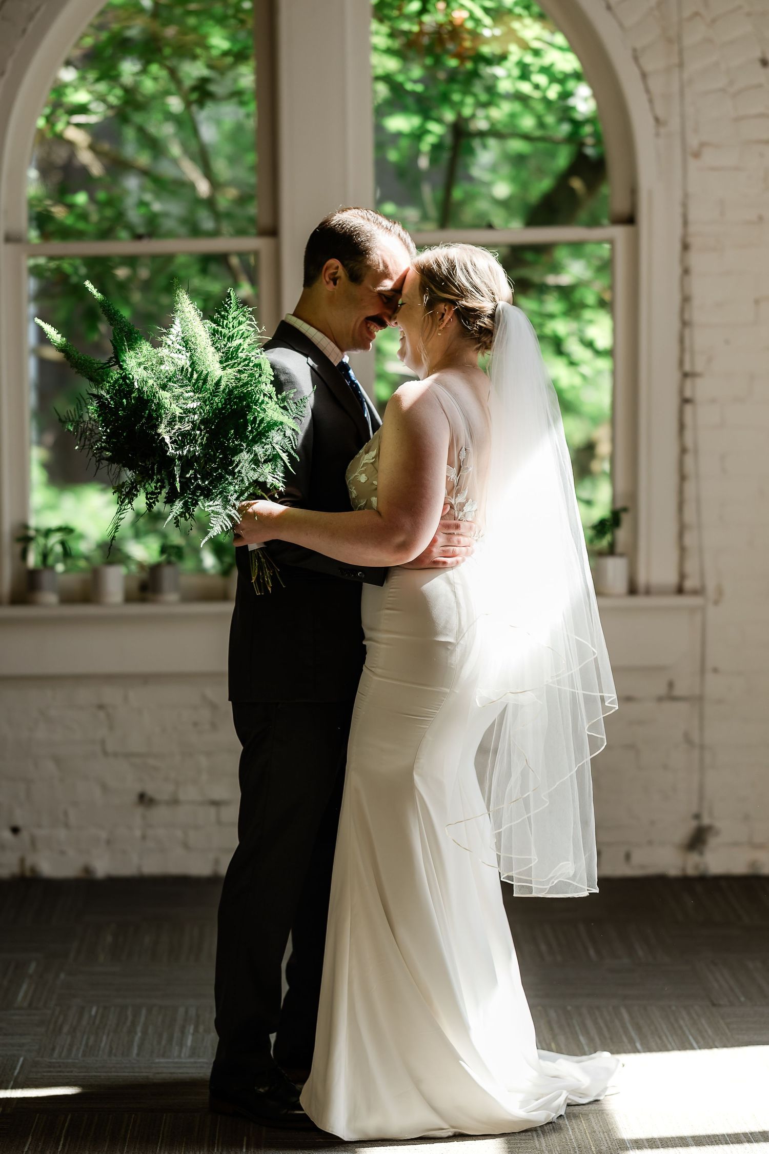 Bride and groom share intimate moment holding green bouquet before arched window.