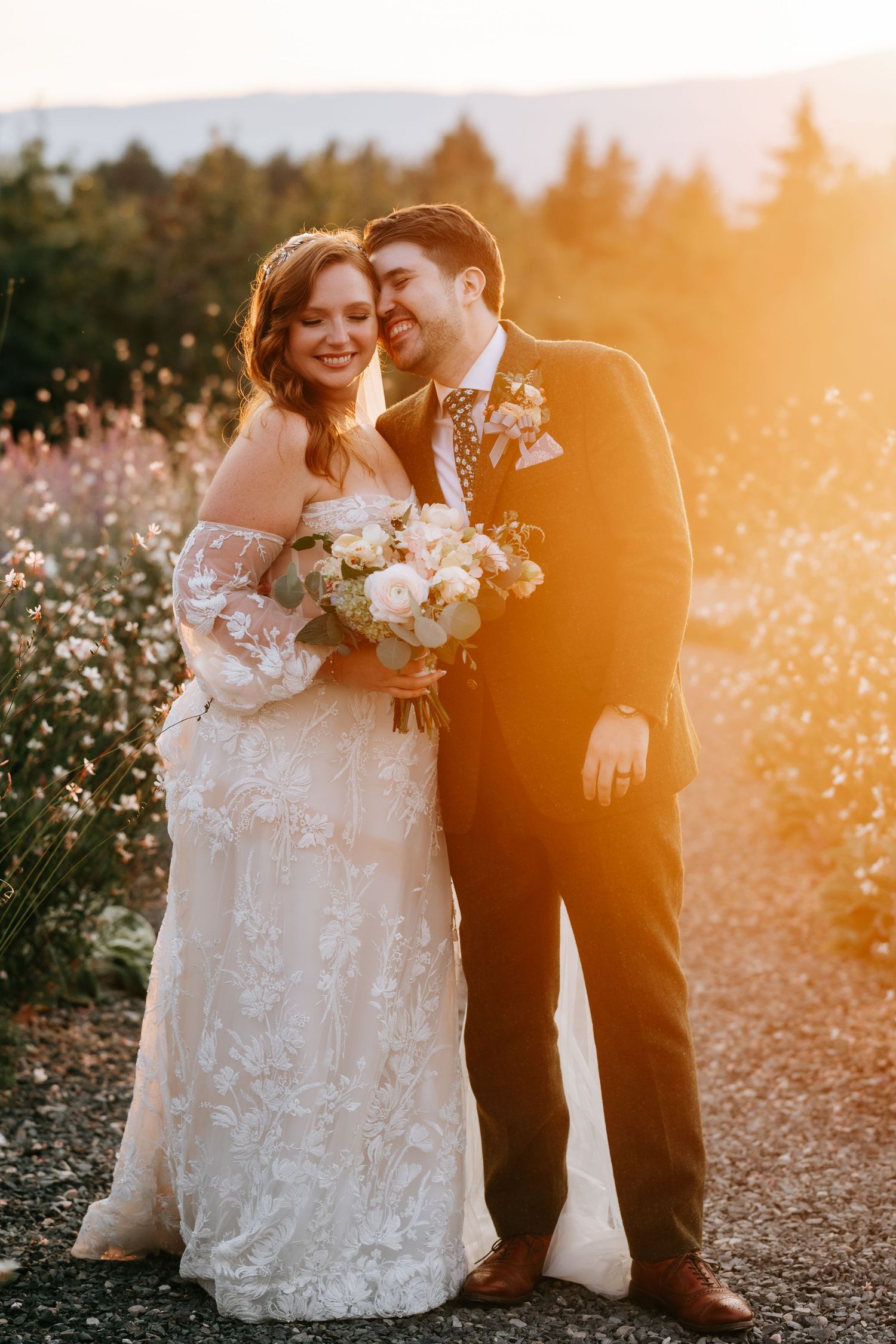 Laughing bride and groom embrace in warm golden sunset light in a blooming garden setting.