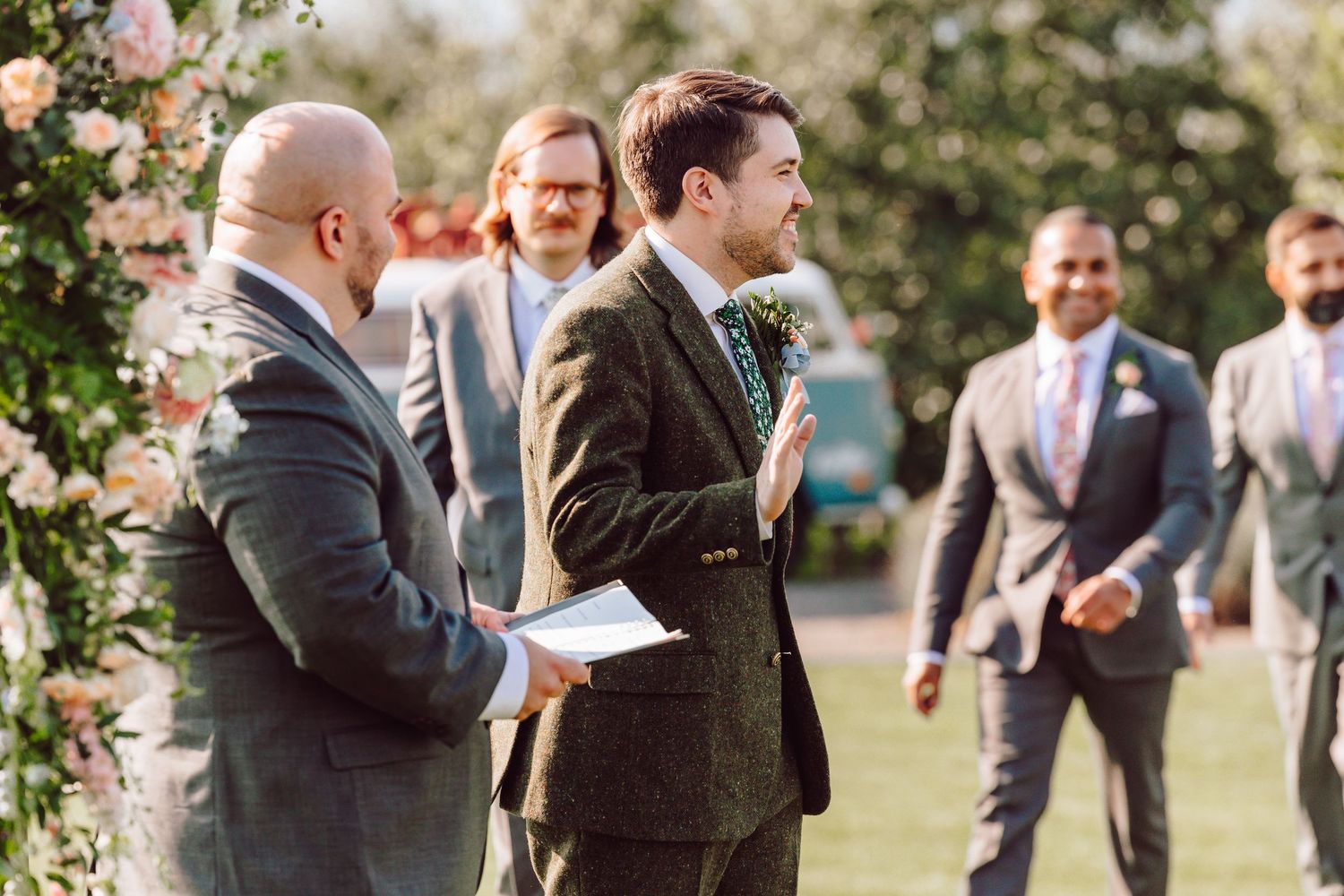 Groom in a green tweed suit listens attentively during an outdoor wedding ceremony.