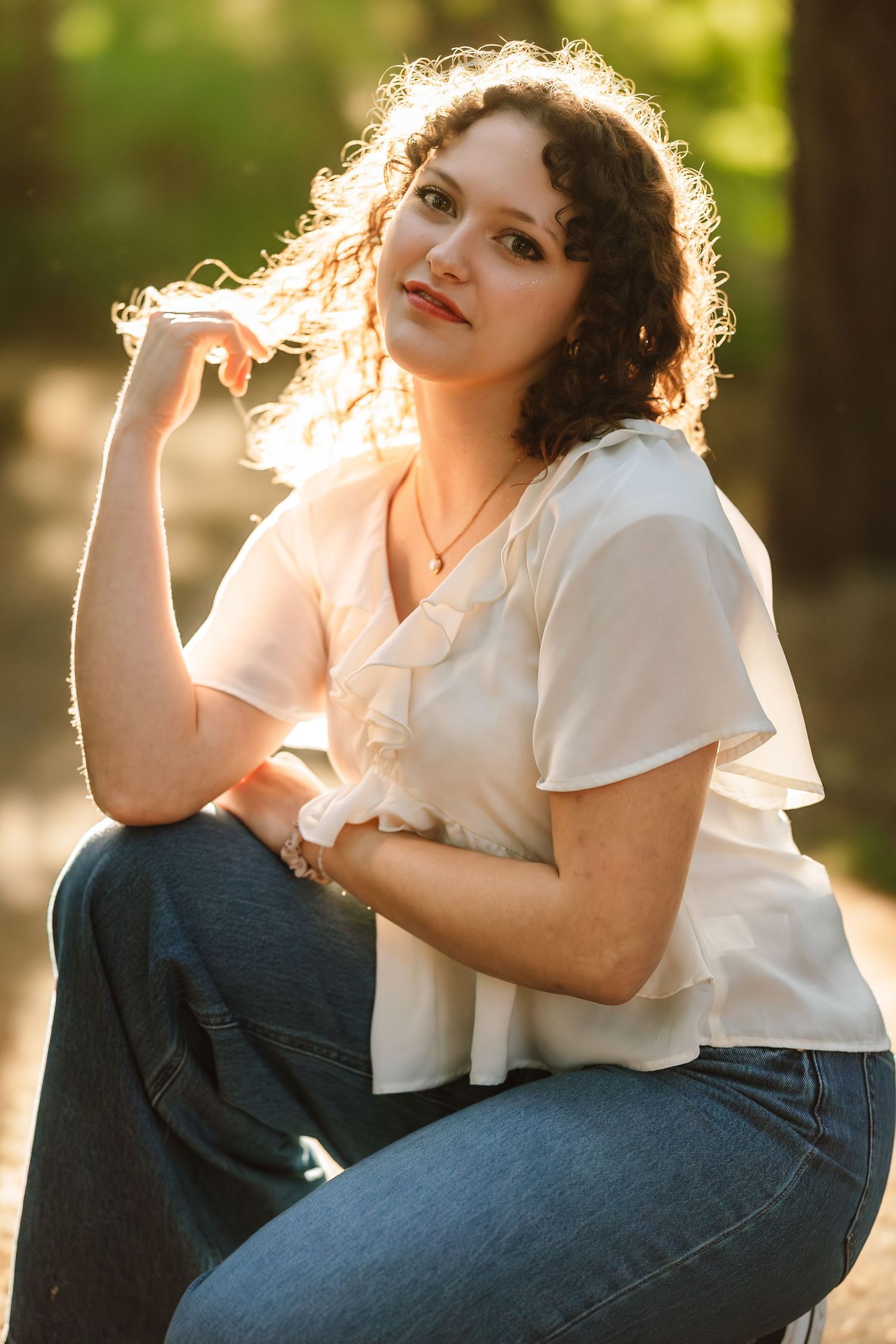 Young woman with curly hair in white blouse and jeans poses outdoors in golden sunlight among trees.