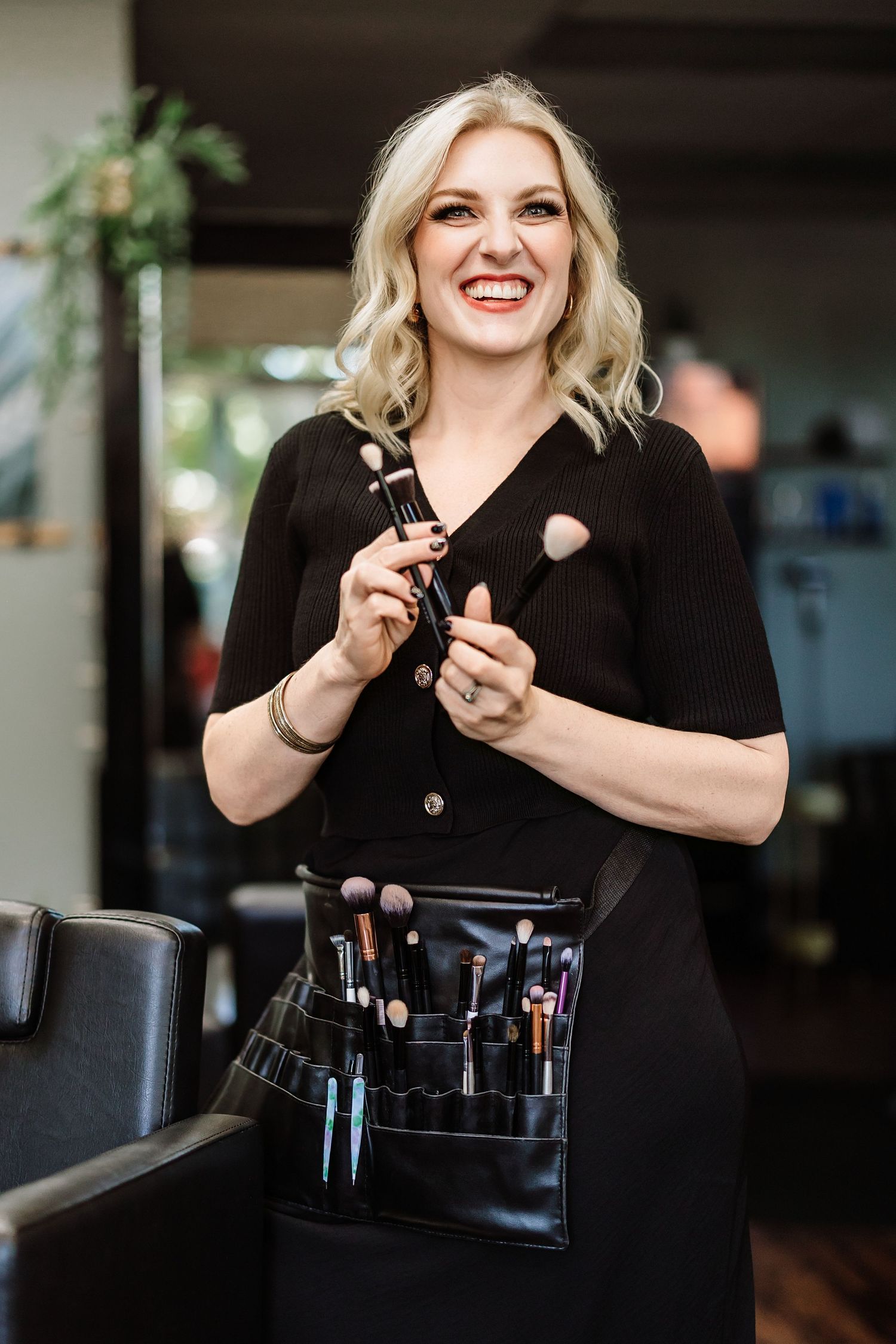 Smiling blonde makeup artist in black dress holding brushes with professional brush belt.