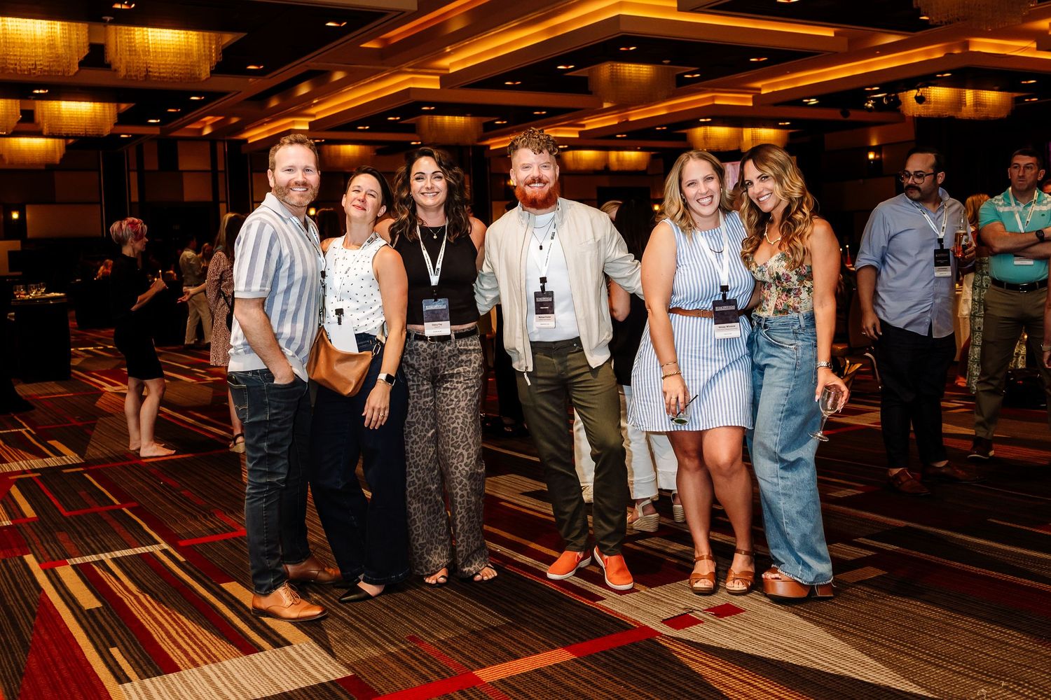 Group of six young professionals smiling together on patterned carpet at a conference event.