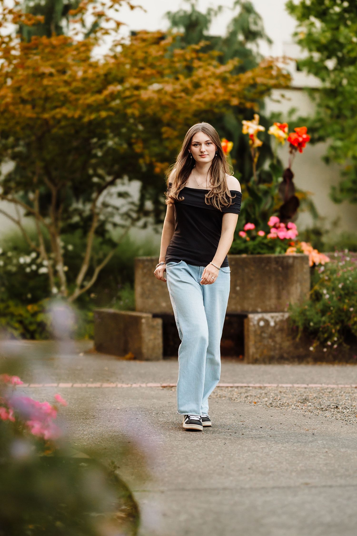 Young woman in black off-shoulder top and light blue jeans walking through a colorful garden path.