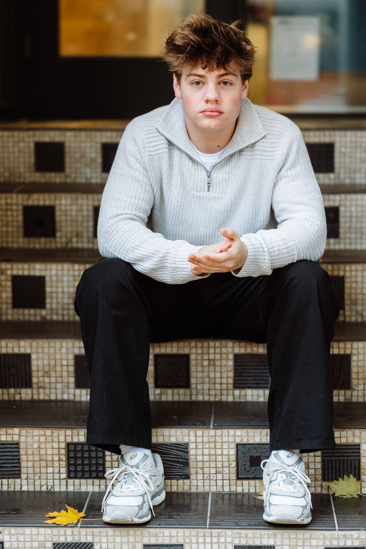 Young man in gray sweater and black pants sitting on decorative tiled steps wearing white sneakers.