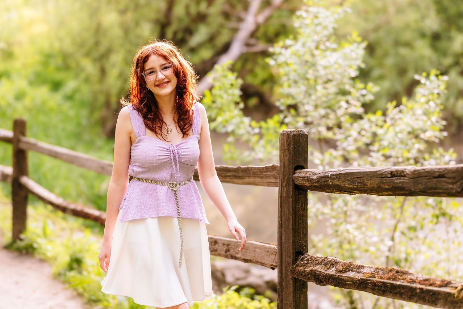 A smiling redhead woman in a lavender top and white skirt leans against a rustic wooden fence outdoors.