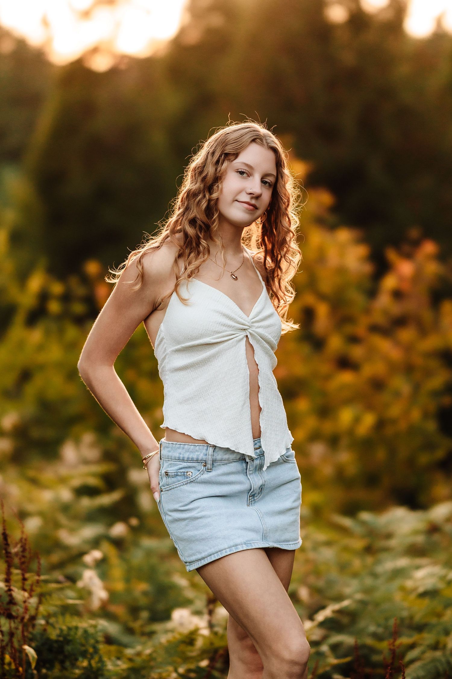 Young woman in white crop top and denim skirt posing outdoors during golden hour sunset.