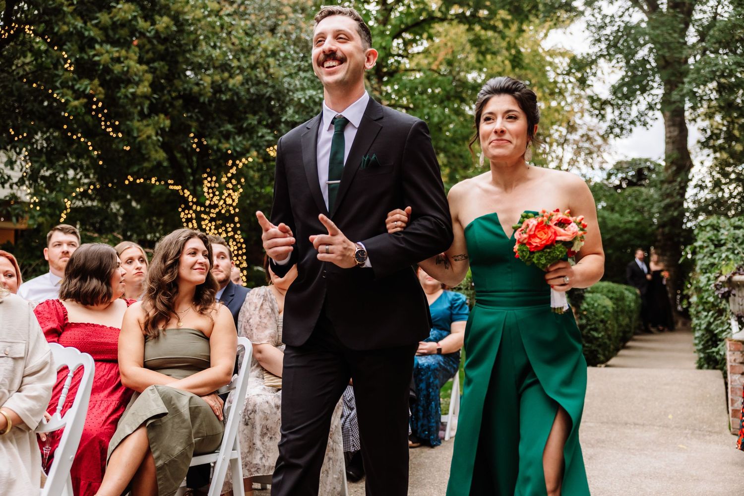 Smiling groomsman escorts bridesmaid in green gown down outdoor wedding ceremony aisle.