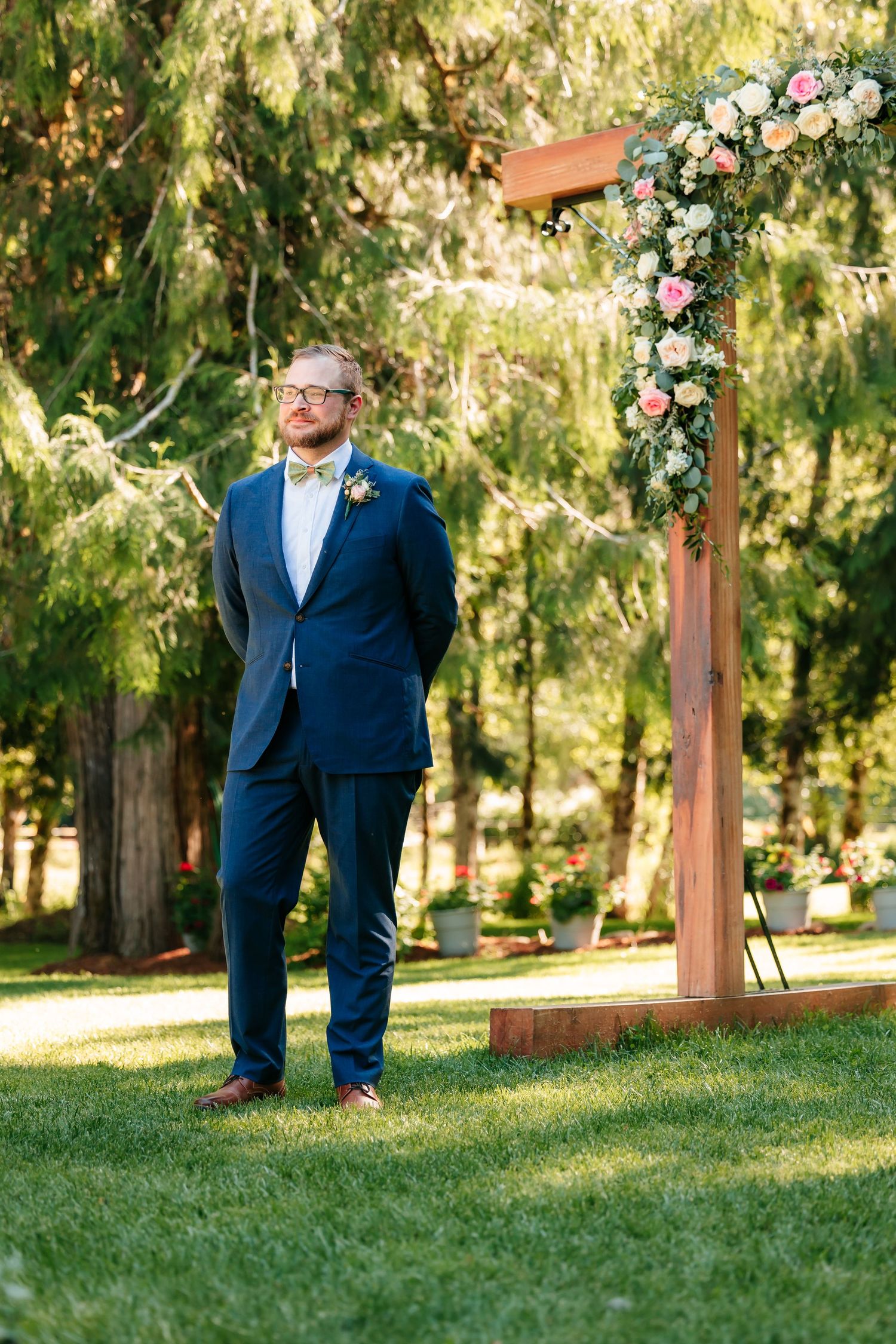 Groom in navy suit with yellow bow tie stands beside floral arch at outdoor wedding ceremony.