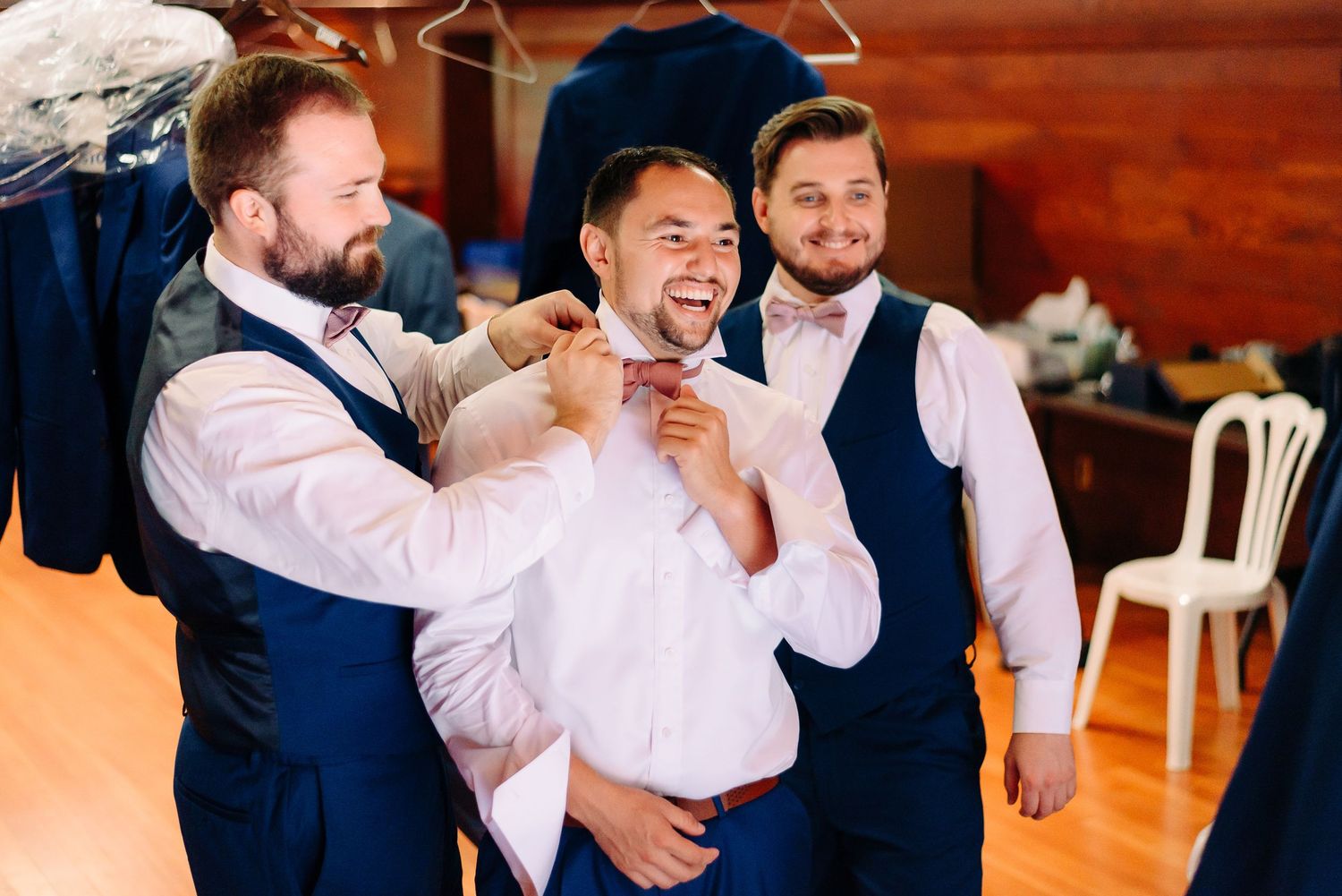 Two groomsmen in blue vests helping the groom adjust his bow tie while laughing together.