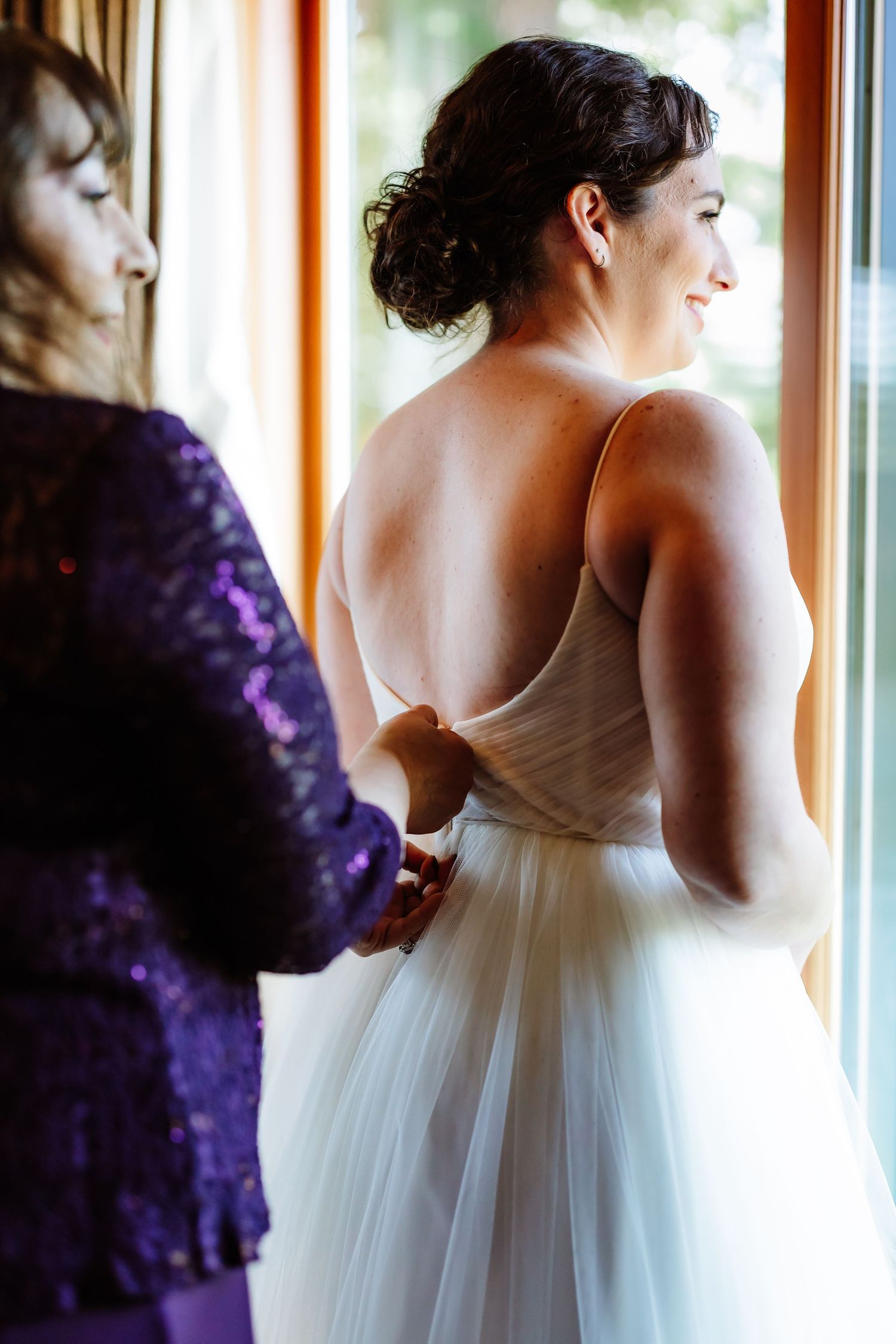 Helper fastens the back of a brides elegant white tulle gown near a sunlit window.