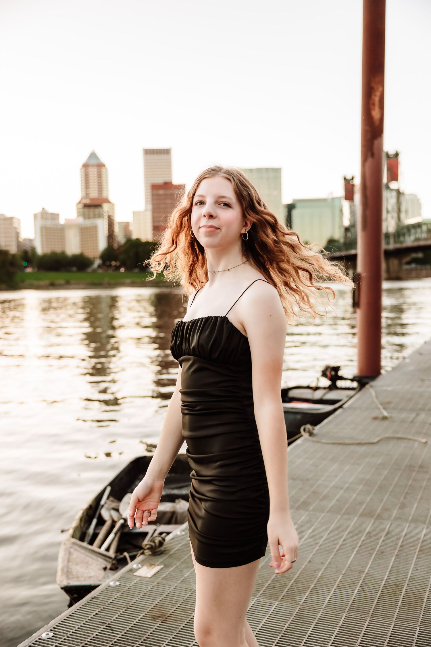 Young woman in black mini dress poses on waterfront dock with city skyline at golden hour.
